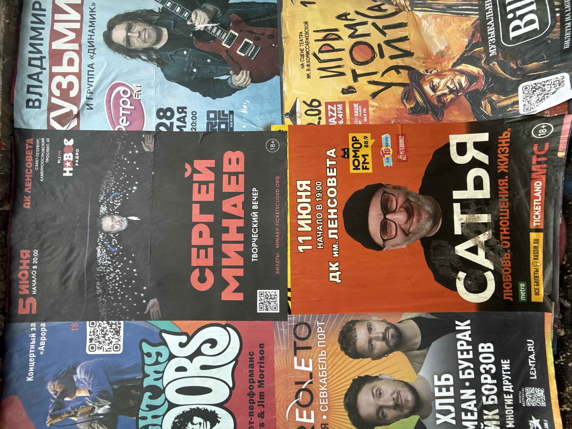 The image shows a collection of event posters for musical acts and performances in Saint Petersburg, Russia. The posters advertise various concerts and shows. Artists such as Vladimir Kuzmin, Sergei Minaev, and the bands The Doors, Khleb, are featured. The posters also mention specific venues such as the "Avrora" Concert Hall, DK im. Lensoveta (Lensoviet Palace of Culture), and Sevkabel Port. Some posters mention radio stations such as Retro FM, Nashe Radio, and Humor FM. There are age restrictions (18+) for some events, and the posters promote ticket sales through websites like Kassir.ru, Ticketland, and Lenta.ru.