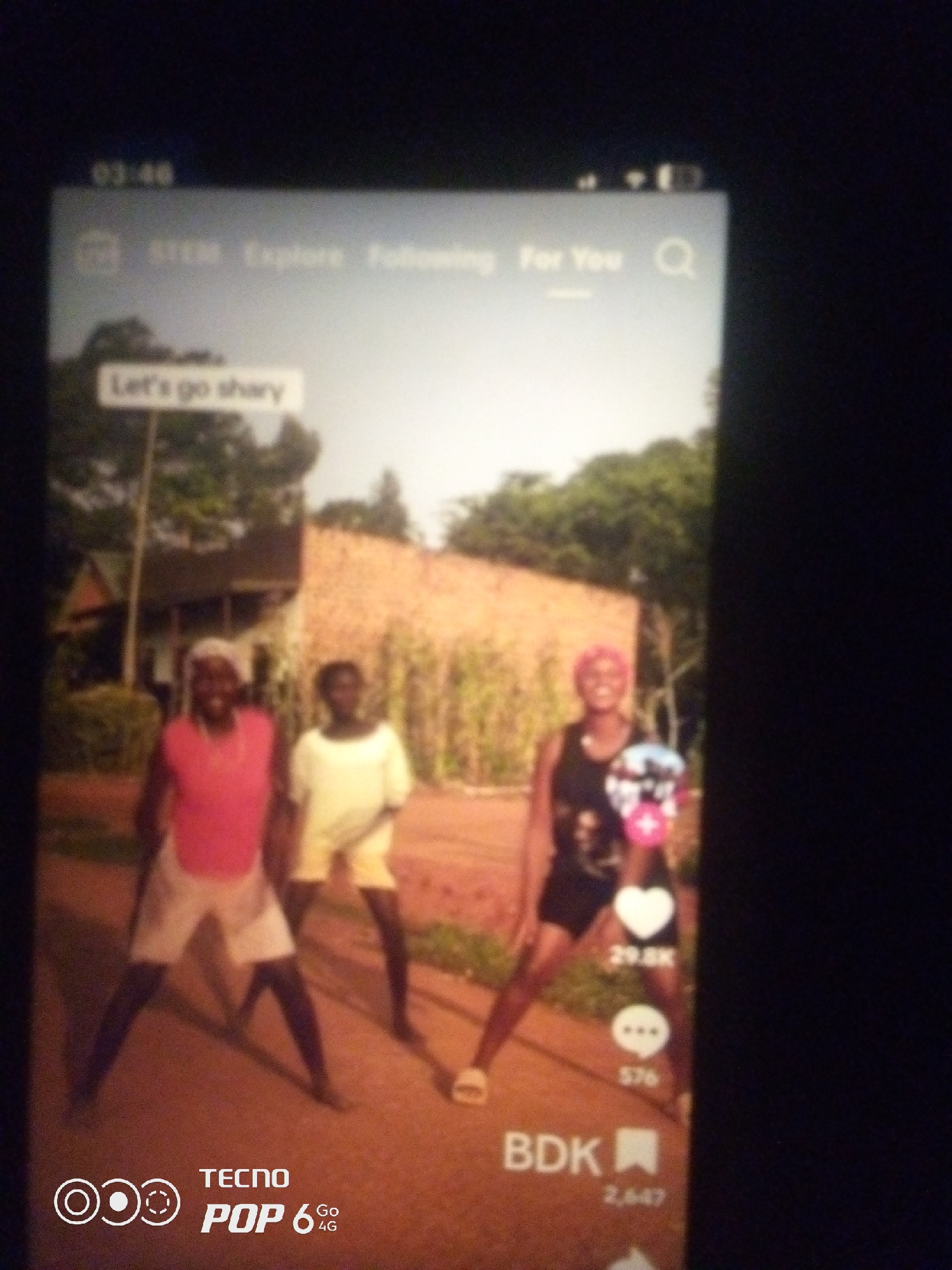 The image shows a screenshot of a social media video, likely from TikTok, overlaid on a dimly lit surface. The video itself depicts three individuals standing outdoors. The background suggests a rural or semi-rural setting with a building with a thatched roof and a cornfield visible. The individuals appear to be young adults, with two of them wearing casual clothing. One person is wearing a pink sleeveless top and white shorts, another is in a white t-shirt and yellow shorts, and the third is in a black tank top and dark shorts with a pink headscarf. Text overlays on the video indicate social media engagement metrics, such as likes and comments, as well as a user handle "BDK". The time stamp "03:46" is visible in the top left corner of the screen. The lower part of the screen shows the TECNO POP 6 4G logo. The overall image quality is poor, with blurriness and low resolution. There is no visual information in the image that can confirm or suggest the location as Howayi, Nigeria.