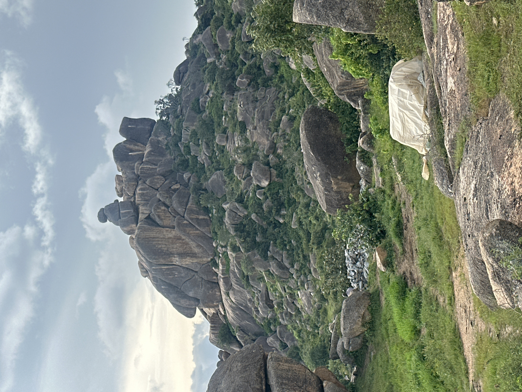 The image depicts a rugged, rocky landscape with a prominent, weathered rock formation dominating the background. The rocks are large and irregular, with some appearing stacked or balanced precariously. Lush green vegetation, including trees and shrubs, covers the slopes and areas between the rocks. In the foreground, the terrain transitions to a grassy area with some scattered rocks and a covered object, possibly a tarped item, placed on the ground. The sky is partly cloudy, suggesting daytime conditions. The scene is indicative of a natural, outdoor environment, likely in a hilly or mountainous region. The location is identified as part of the city of Jos, Nigeria, a region known for its unique geological formations and rocky outcrops.