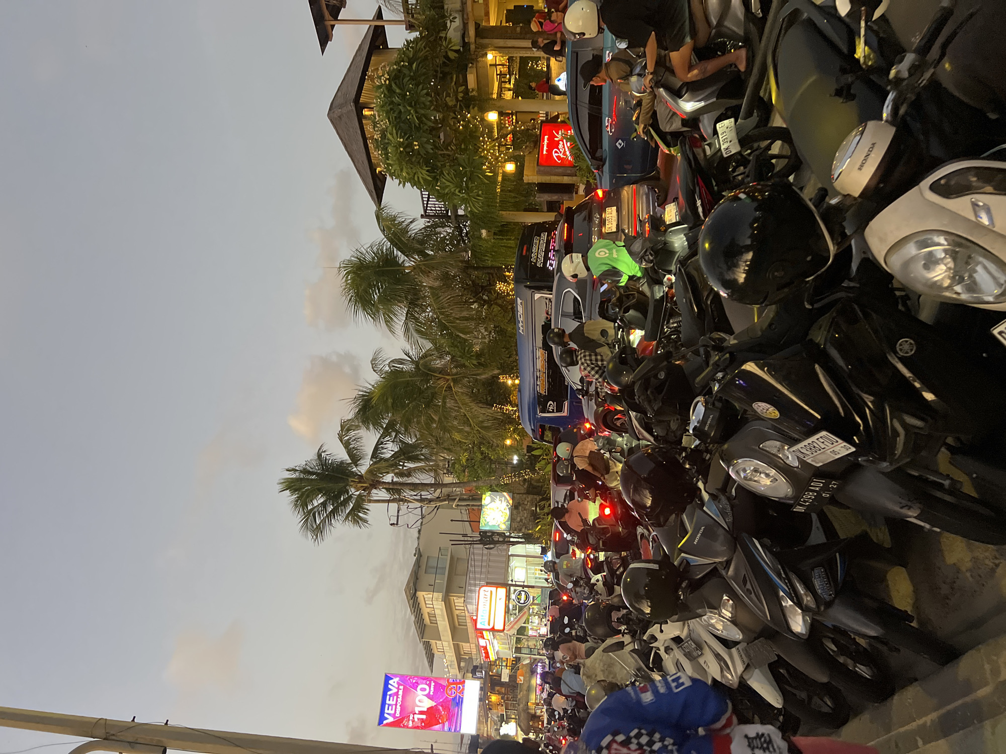 The image depicts a crowded street scene in Kuta, Indonesia, at dusk. A significant number of motorcycles are parked and in motion, indicating heavy traffic. Several people are visible, some riding motorcycles, others walking or standing. Buildings line the street, including commercial establishments like "Alfamart" and "Veeva Disposable" with prominent signage. Palm trees are scattered throughout the scene, suggesting a tropical or subtropical climate. The overall atmosphere is one of bustling activity, typical of a busy urban area in Indonesia.