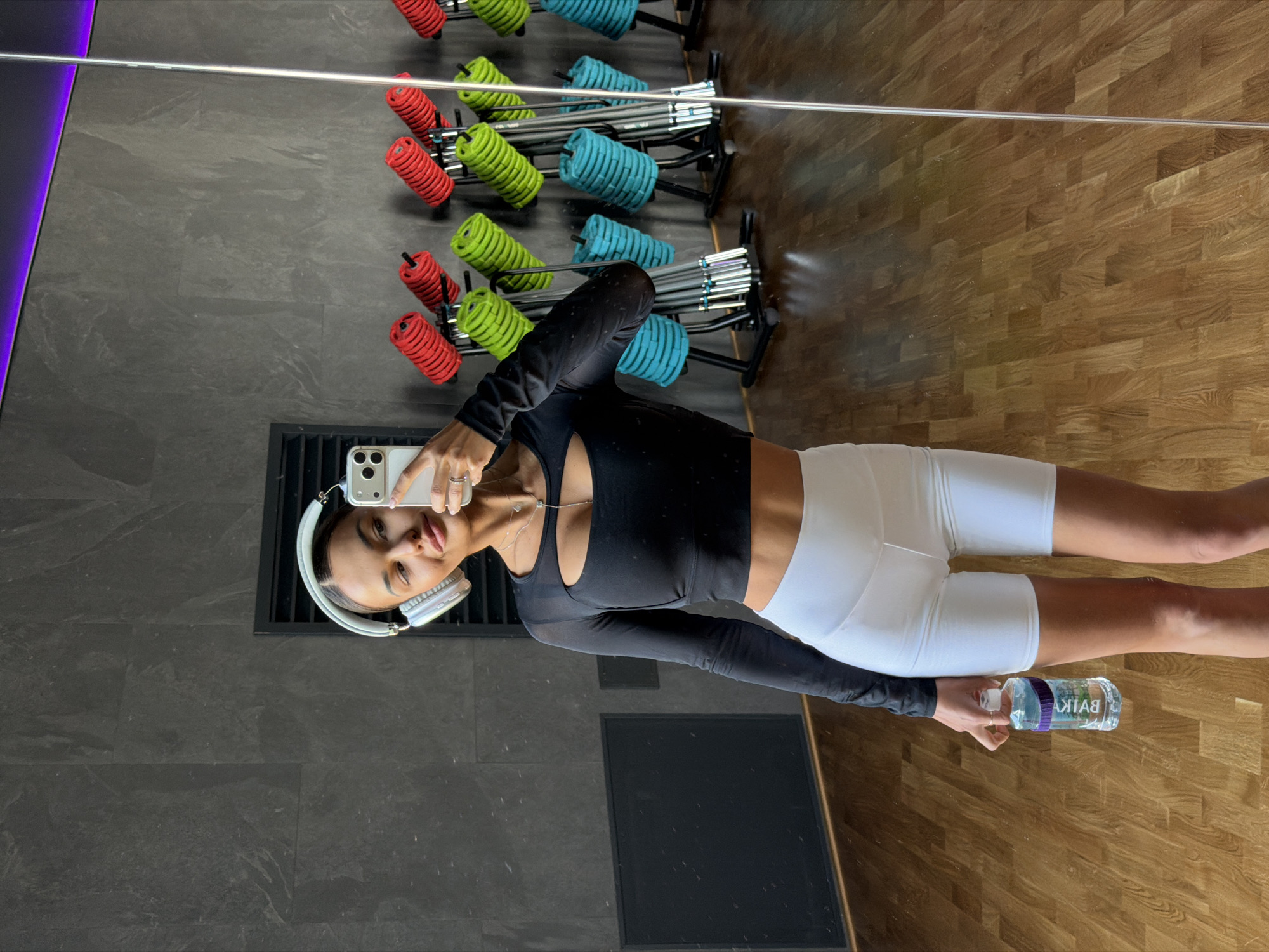A woman is standing in a gym, taking a selfie in a mirror. She is wearing a black long-sleeved crop top and white athletic shorts. She is holding a water bottle in one hand and her phone in the other, wearing headphones around her neck. The gym has gray tiled floors on one side and wooden floors on the other. There are rows of colored weights (red, green, and teal) on racks behind her. The location cannot be confirmed as being in Krasnoyarsk, Russia.
