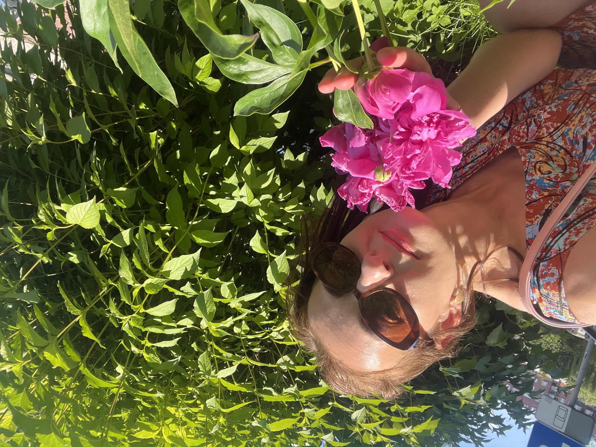 This is a close-up, upside-down, outdoor shot of a person holding a bouquet of bright pink peonies. The person is wearing sunglasses with brown lenses and a colorful, abstract-patterned top. Their lips are slightly parted, and their eyes are closed. The background is dominated by lush green foliage, suggesting a garden or park setting. The lighting indicates it is daytime, likely sunny, casting shadows and highlighting the vibrant colors of the flowers and leaves. The overall mood seems relaxed and joyful, with the person enjoying the beauty of the peonies in a natural environment. There is a faint suggestion of buildings in the far background, hinting at an urban or suburban location. A blue street sign with white text is partially visible in the lower right corner.