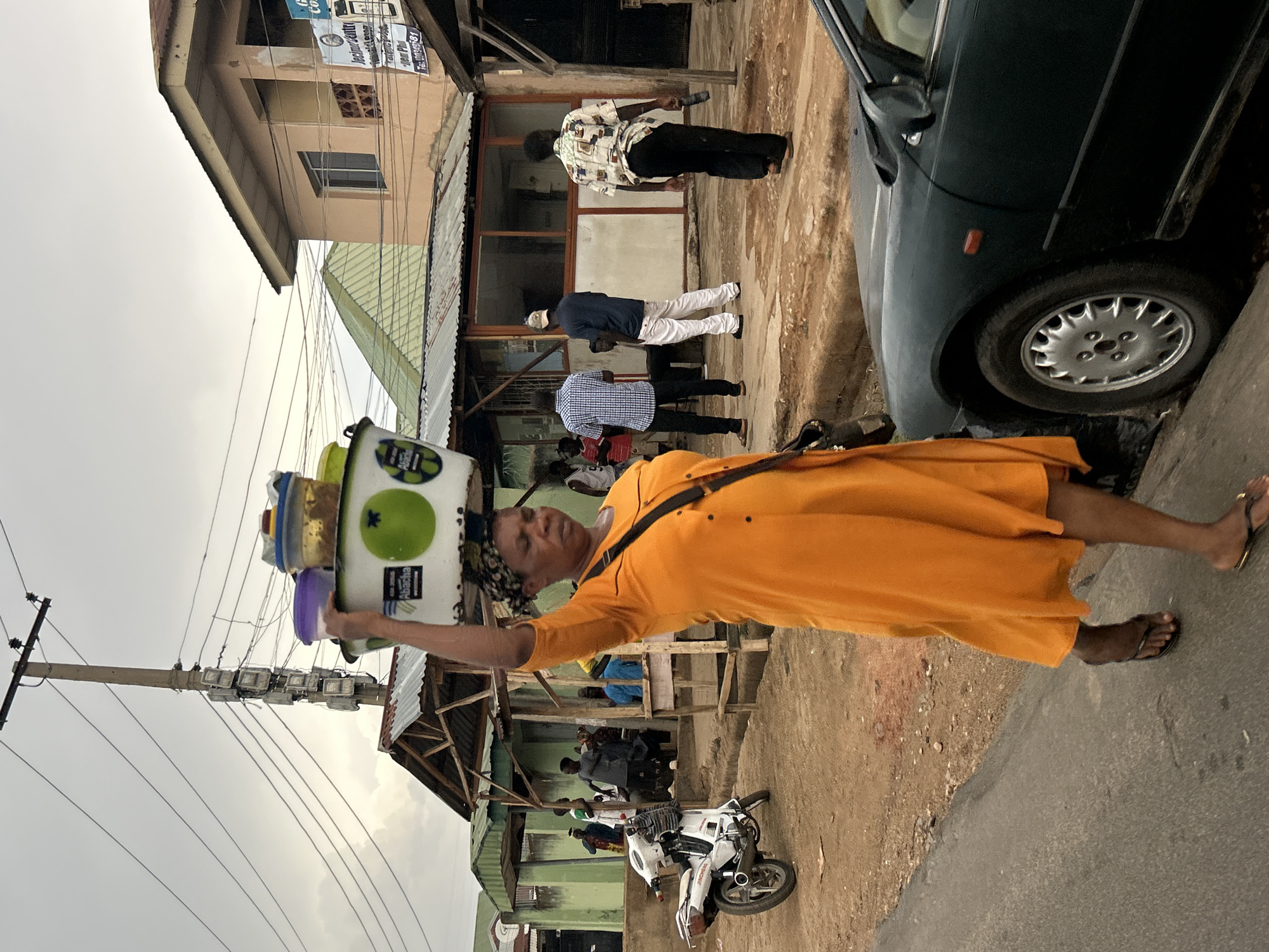 A street scene in Jos, Nigeria, shows a woman wearing an orange dress and sandals carrying containers on her head. The woman is walking along a street with a car parked nearby. In the background, there are buildings and people standing under a covered area, and a motorcycle parked. Overhead power lines and a utility pole are visible. The sky is overcast.