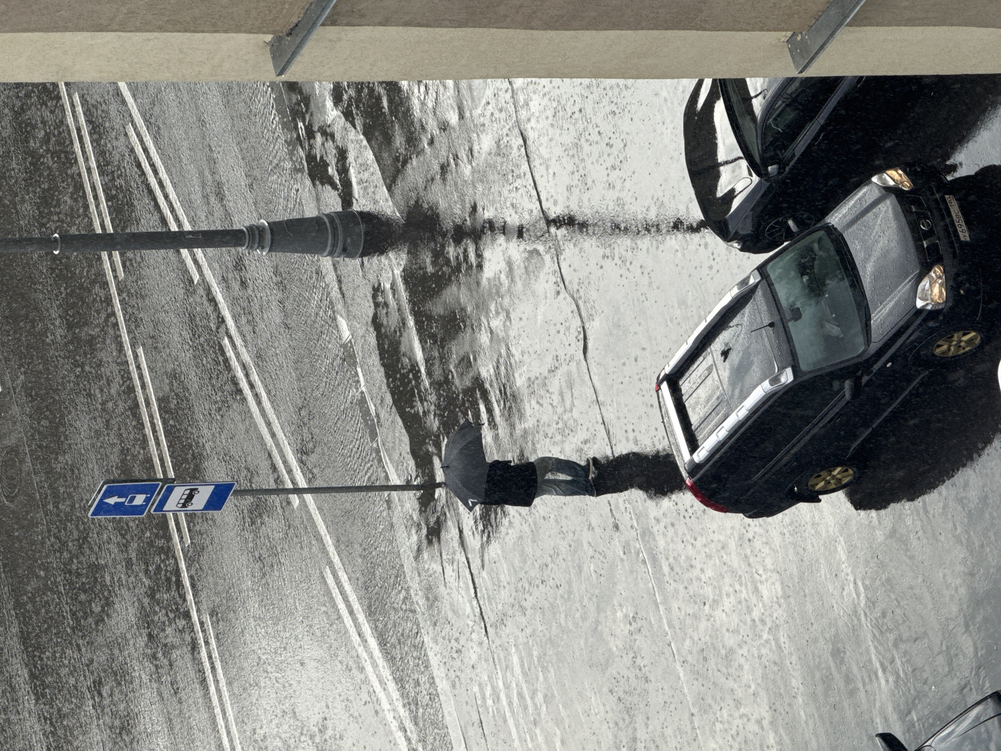 An overhead shot captures a rainy street scene. A person, holding a dark umbrella, stands on the wet asphalt, their reflection visible in the puddles. The person is wearing jeans and a dark jacket. Adjacent to them, a signpost displays two blue rectangular signs with white pictograms. The left sign indicates a leftward arrow, while the right sign depicts a bus. Further into the scene, two dark-colored cars are parked on the wet road. The car on the right is partially visible, with its rear bumper and taillight illuminated. The car on the left has its driver's side door open. The road is marked with white lines, suggesting lanes of traffic. The overall atmosphere is gloomy, indicated by the overcast sky and the rain. The scene is likely taking place during the daytime. The license plate of the car on the right is partially visible and appears to read "6958BC 190".