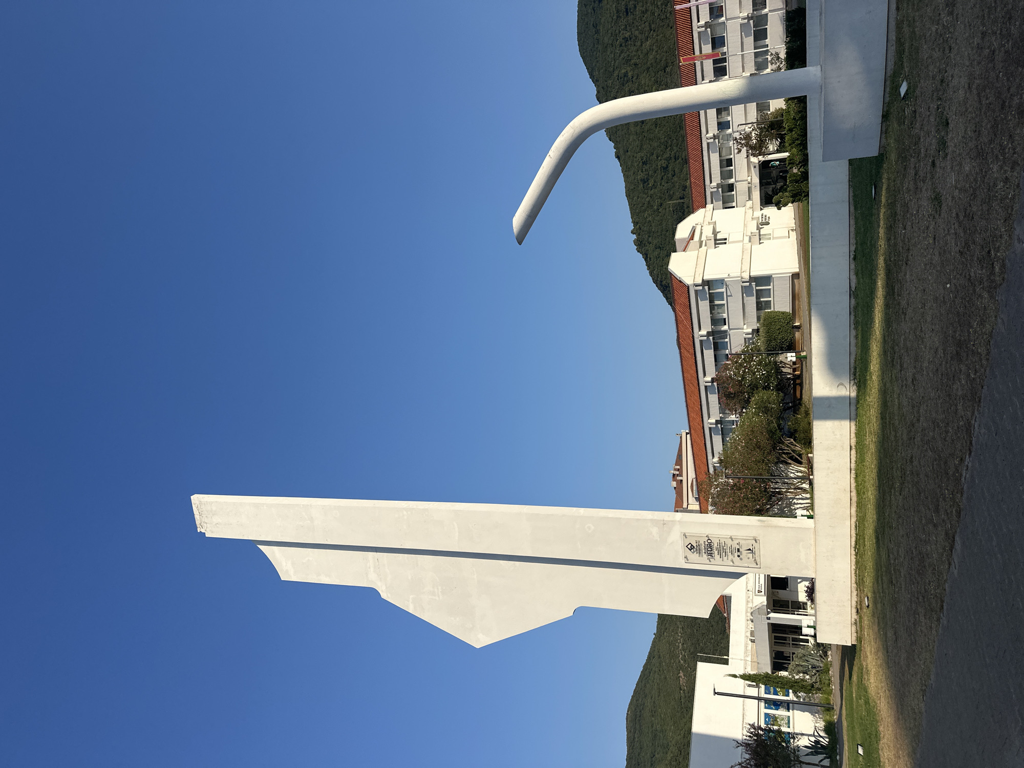 The image depicts a large, abstract white sculpture with a sharp, angular form in the foreground. To the right of this sculpture, a curved white structure, resembling a lamppost or a modern art installation, is visible. In the background, there is a white building with a red-tiled roof, characteristic of Mediterranean architecture. The building appears to be a school or administrative complex, with multiple windows and a sign that is partially legible. Green, tree-covered hills rise behind the building, and the sky is a clear, bright blue. The ground in front of the sculpture is a patchy, dry lawn. The overall scene suggests an outdoor public space, possibly a plaza or park, in a coastal town given the vegetation and architectural style. The sculpture itself has a plaque on its base. Based on context from similar images, this is likely the monument dedicated to the partisans of the Second World War, located in Budva, Montenegro, specifically near the elementary school "Veljko Drobnjakovic".