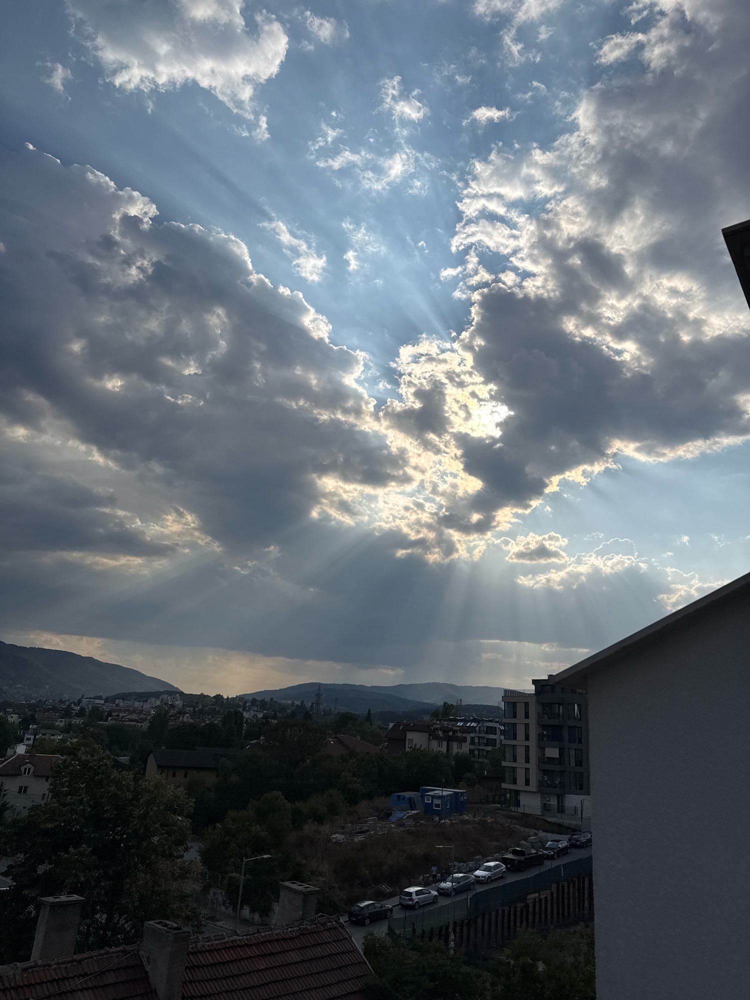 The image captures a city view, likely from an elevated position in Sofia, Bulgaria, during what appears to be late afternoon or early evening. The sky is dramatic, with a mix of dark, heavy clouds and brighter, sunlit patches. Rays of sunlight pierce through the clouds, creating a crepuscular effect, illuminating parts of the sky and casting shadows over the landscape below.
In the foreground, the edge of a building's roof and wall is visible on the right side. Below this, the scene opens up to a residential area. Houses with tiled roofs and trees are interspersed with modern apartment buildings. A street lined with parked cars is visible, with construction fencing and materials suggesting ongoing development or renovation. In the distance, rolling hills or mountains form the horizon, shrouded in a hazy, muted light. The overall impression is one of urban life under a dynamic and visually striking sky.