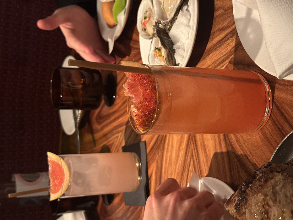 The image captures a dimly lit, intimate setting, likely a high-end restaurant or bar in London. The scene is dominated by a polished wooden table with a distinctive striped grain pattern.
In the foreground and center, two tall, narrow glasses contain vibrant cocktails. The cocktail on the right is a reddish-orange hue, garnished with a segment of citrus (likely grapefruit) that has a vivid red, granular rim, possibly chili salt or spiced sugar. A thin, light-colored straw extends from the drink. To its left, another similar glass holds a lighter, cloudier pink or off-white beverage, also adorned with a fresh grapefruit slice and a straw. This glass rests on a dark, rectangular coaster.
Various food items are arranged on the table. In the upper center, a white oval plate holds several fresh oysters nestled on a bed of ice. Above this, another small white oval dish contains a small, golden-brown item, possibly an appetizer or fritter, garnished with a bright green leaf. A dark brown glass tumbler is visible behind the lighter cocktail. In the bottom right corner, a piece of crusty bread, possibly sourdough, is partially visible. A folded white napkin is also seen towards the upper right.
Two sets of hands indicate the presence of at least two people dining. One hand is in the upper left quadrant, reaching towards the plate with the small appetizer. Another hand is visible in the bottom right, near the bread, appearing to hold or interact with a small white dish or napkin. The overall ambiance suggests a sophisticated dining experience, likely during the evening due to the subdued lighting. No discernible text is visible in the image.