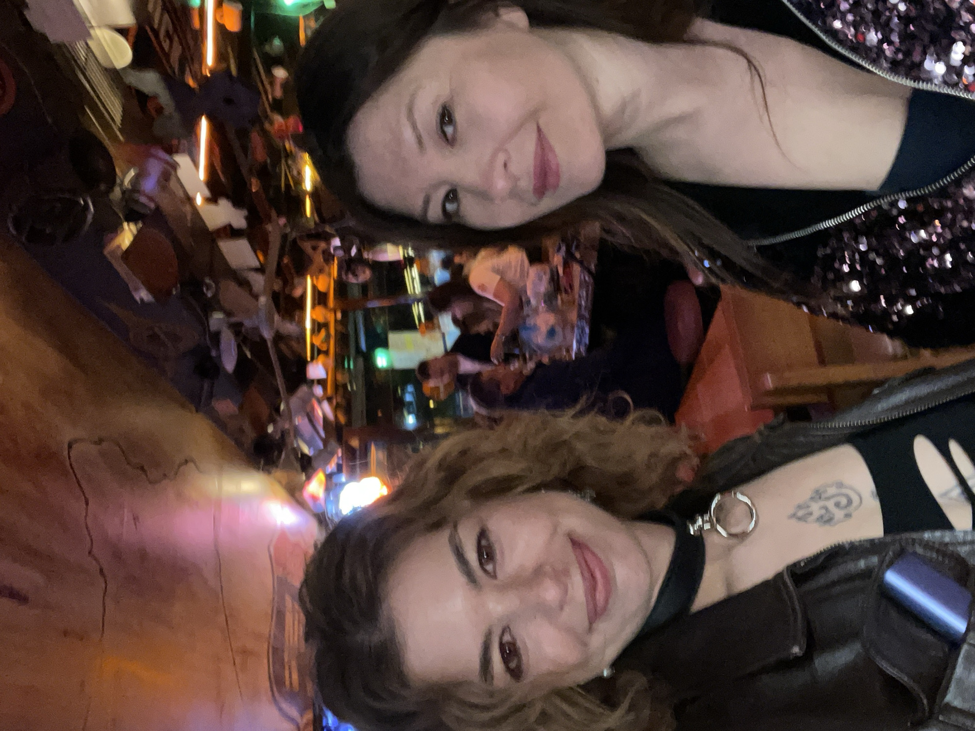 Two women are in a bar or restaurant in Budva, Montenegro. The woman at the top is wearing a sequined jacket. The other woman has curly hair and a leather jacket. She also has a tattoo and a choker.