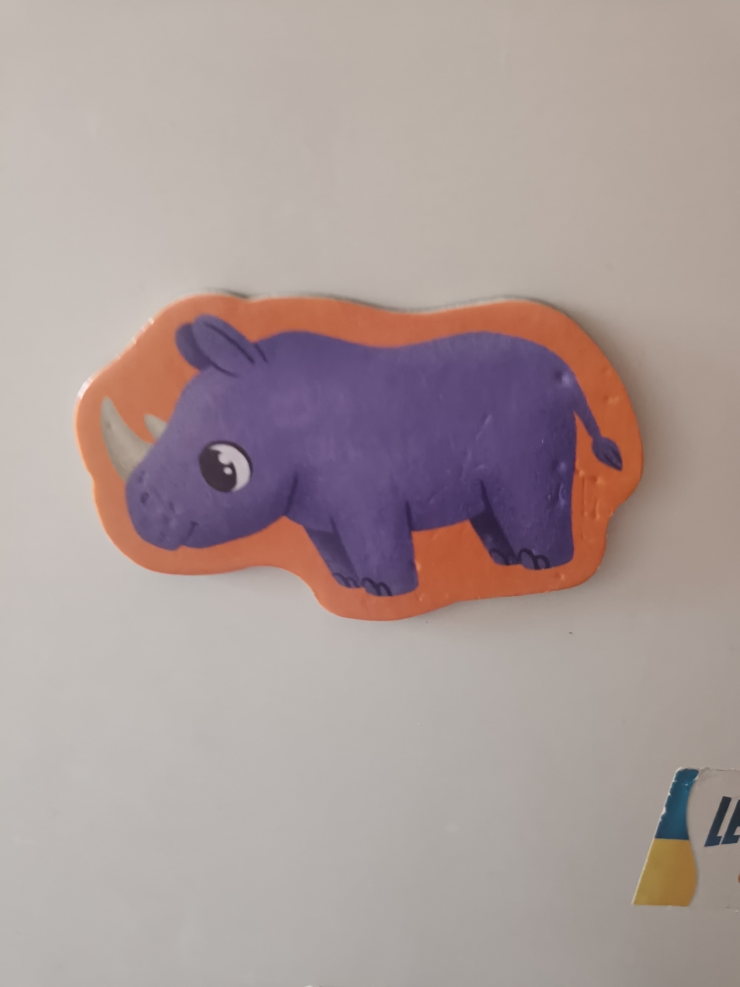 The image shows a purple cartoon rhinoceros sticker on an orange background, adhered to a white surface, likely a refrigerator door, in an indoor setting. The rhinoceros is depicted in profile, facing left, with a large eye, a single horn, and two front legs visible. The sticker has a wavy, irregular outline. In the lower right corner of the image, a portion of a blue and yellow sticker with the partial word "LE" is visible. The background is a plain, light-colored surface, suggesting an interior wall or appliance. There is no visible indication of the city of Odesa, Ukraine, or any other specific location.