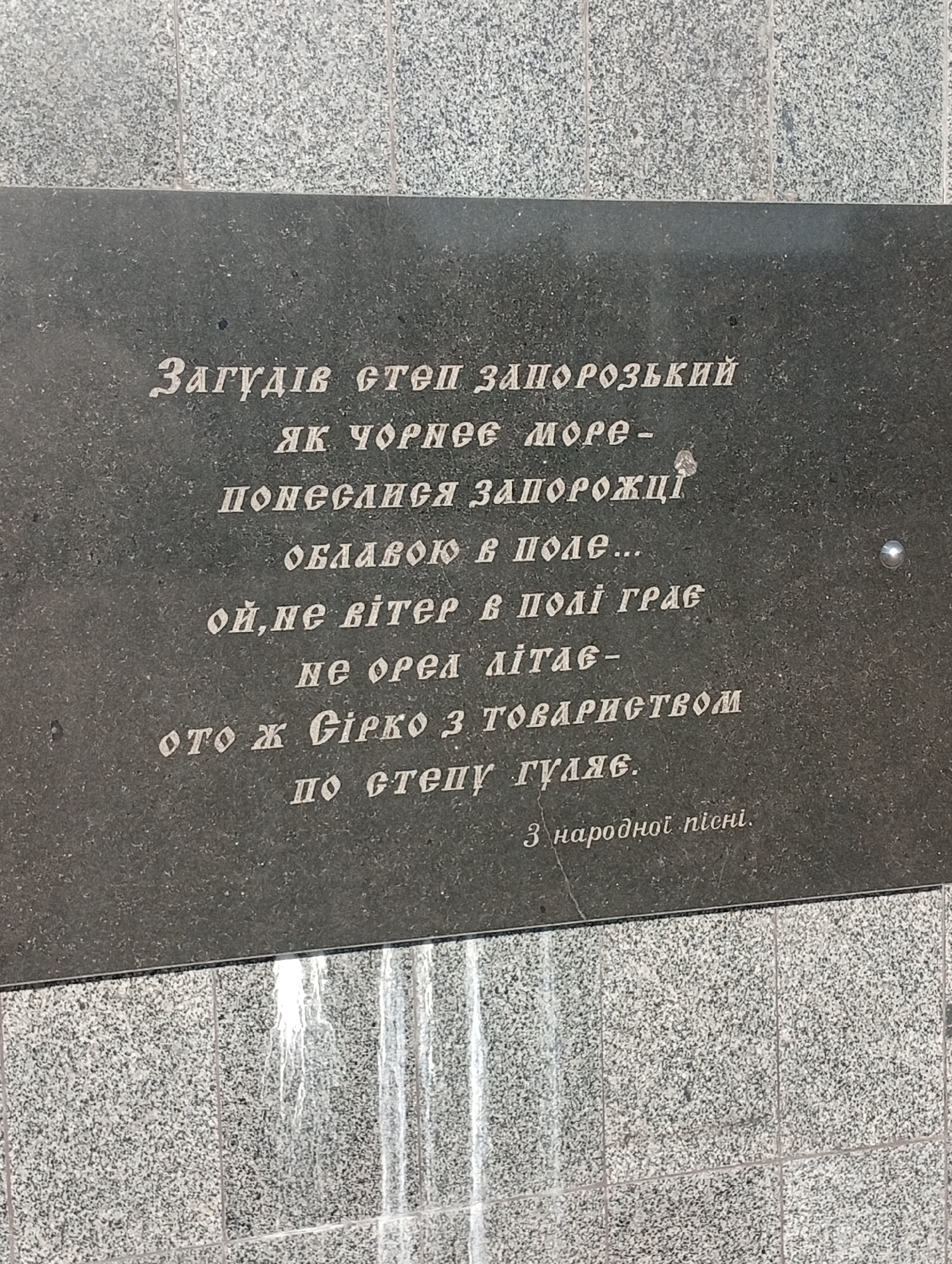 This is a close-up, eye-level photograph of an engraved stone plaque, likely a monument or memorial, set against a textured granite wall. The plaque is dark and rectangular, with white text inscribed on its surface. The text appears to be in Cyrillic script and is written in several lines, forming a poem or inscription. The bottom of the image shows a lighter-colored granite surface, possibly the base of the monument, with vertical lines suggesting it's made of multiple tiles or sections. The lighting suggests daytime, but there are no strong shadows to indicate a specific time. The overall mood is solemn and commemorative, appropriate for a public inscription. The text translates to a snippet of a Ukrainian folk song, speaking of Zaporozhian Cossacks wandering the steppe like a storm. The inscription is located in Pokrov, Ukraine.