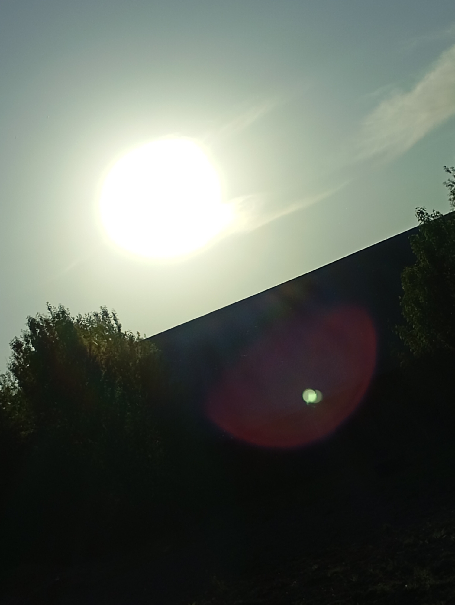 The image shows a bright sun in a pale sky, positioned above a dark, silhouetted landscape. The landscape appears to include trees and a dark, sloping hill or embankment. A red lens flare with a small green reflection is present in the lower right portion of the image. The location is listed as Unknown, Unknown.