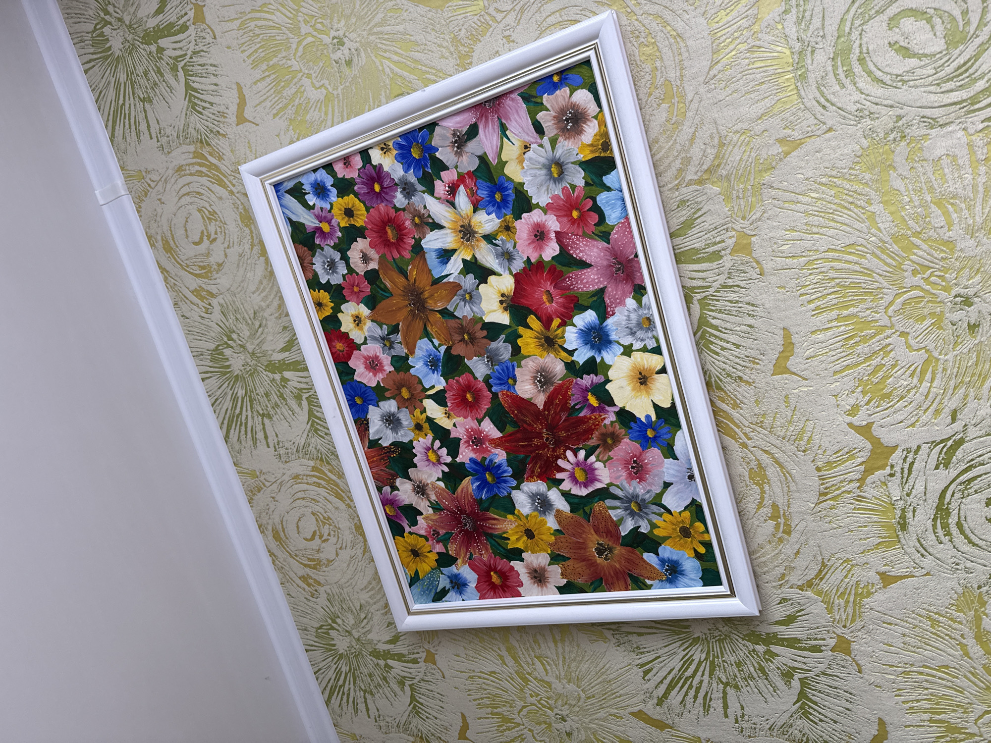 A framed painting of a colorful floral arrangement hangs on a wall in Krasnogorskoye, Russia. The painting is rectangular with a white frame and depicts a dense collection of various flowers in a variety of colors including red, pink, blue, yellow, orange, and white. The flowers are rendered in a detailed, realistic style. The wall behind the painting has a textured wallpaper with a floral pattern in shades of gold and cream. The room appears to be well-lit, with a white ceiling molding visible at the top of the image. There are no people or animals present in the scene. The overall impression is one of domestic decoration.