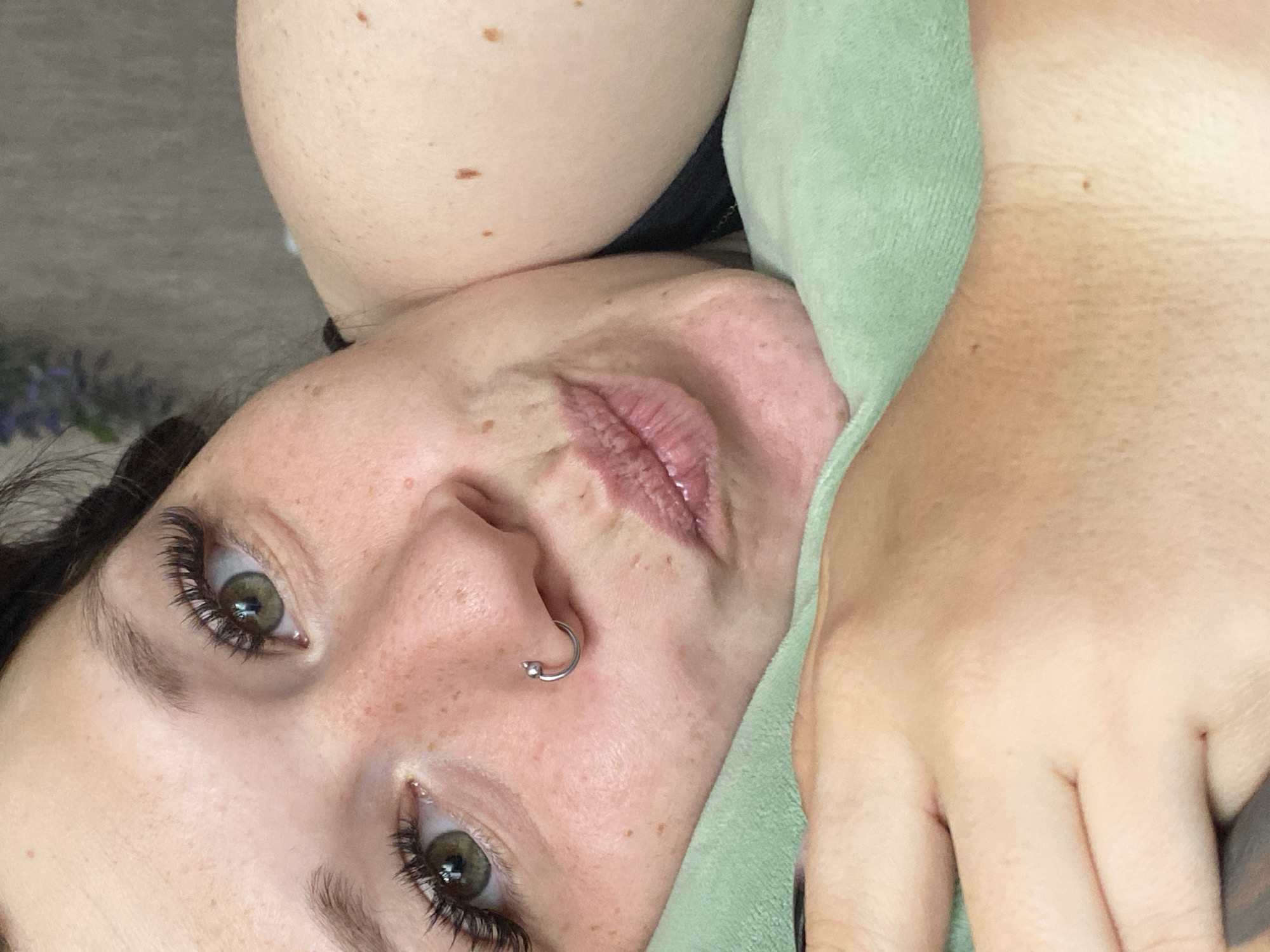 The image is a close-up selfie of a young woman with green eyes and brown hair, wearing false eyelashes and a silver hoop nose ring. She is lying on a light green plush surface, possibly a pillow or blanket, with her head resting on it. Her mouth is slightly puckered as if to give a kiss. Freckles are visible on her cheeks and nose. The background is a blurry, gray, textured surface. The lighting suggests an indoor setting. There is no indication of the specific location within the city of Chaikovsky, Russia.