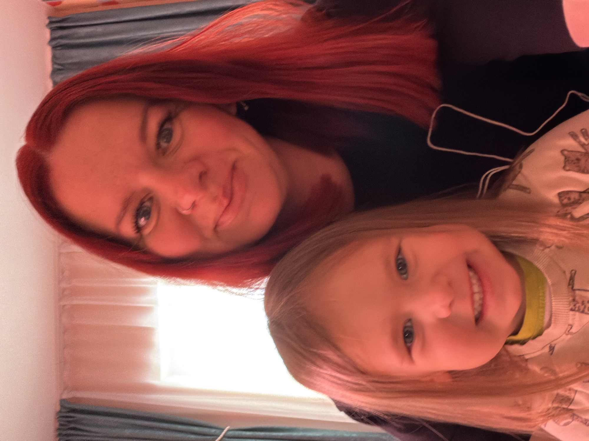 An overhead, slightly rotated photograph captures a candid, smiling moment between an adult woman and a young child, likely a mother and daughter, in an indoor setting.
The adult woman, positioned in the upper left of the frame (when correctly oriented), has striking, vibrant red hair that cascades down her left side. Her light-colored eyes (appearing blue or grey) are visible, and she has subtle eye makeup. She wears a faint smile, and a small nose ring is visible on her right nostril. She is dressed in a dark-colored top, possibly black, with a light-colored trim or design visible on the right side near her shoulder.
To the lower right of the woman is a young child, likely a girl, with blonde hair and bright, light-colored eyes. The child is looking directly at the camera with a wide, joyful, open-mouthed smile, revealing her teeth. She is wearing a light-colored long-sleeved top adorned with a pattern of what appear to be cartoon animals or bears. Underneath her main top, a bright lime green or yellow collar is visible around her neck.
The composition suggests a close-up selfie or a photo taken from above, focusing on the faces of the two individuals. The background is out of focus, indicating a shallow depth of field. On the left side of the frame (when oriented correctly), a bright source of light, likely a window, is visible, partially obscured by sheer white curtains or blinds. Below this, there's a darker, possibly textured, area. The overall lighting is soft and warm, illuminating their faces. Both individuals convey happiness and affection through their expressions. There is no visible text in the image.