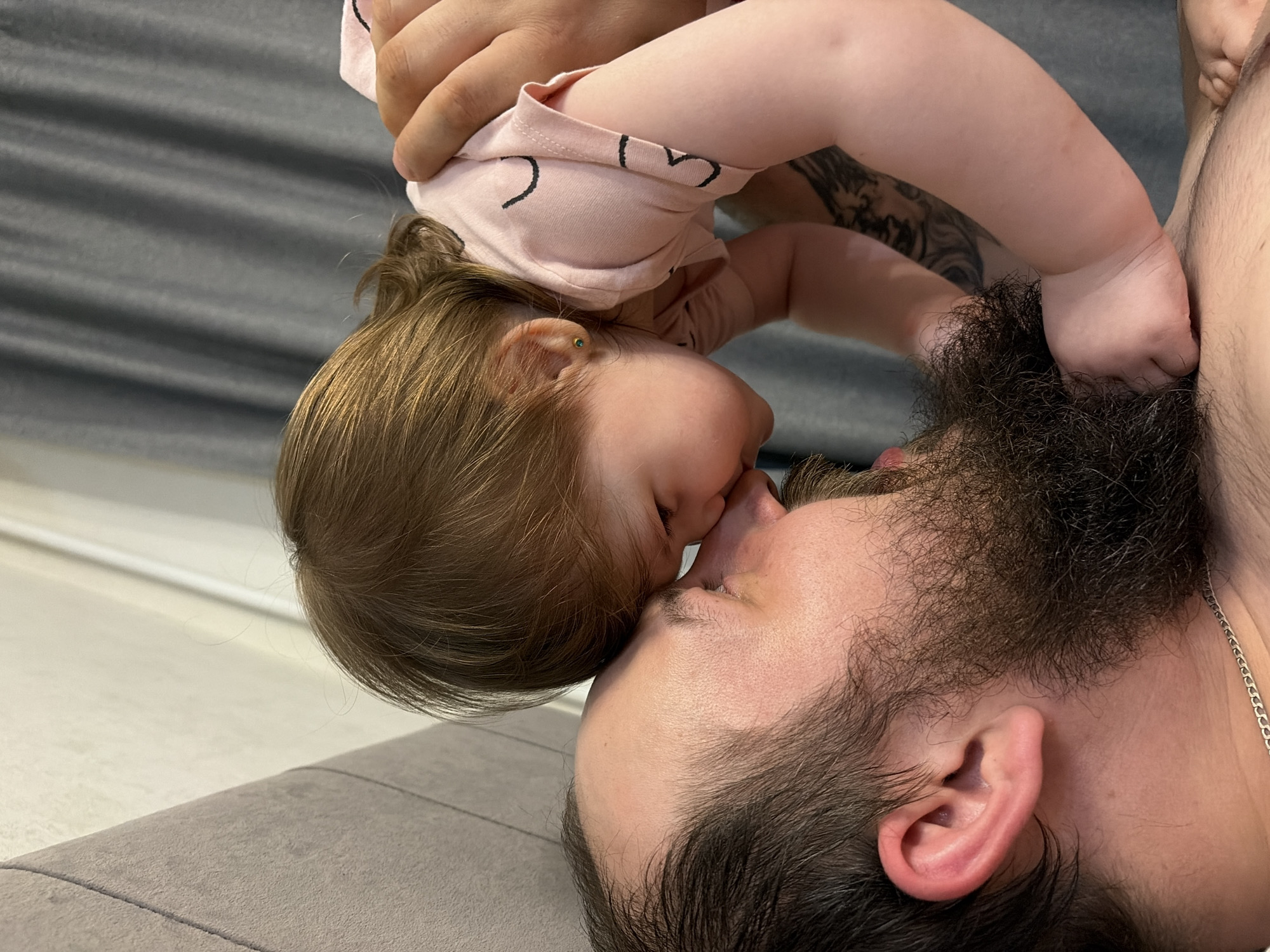 The image shows a close-up of a man lying on a gray sofa with a baby being held above him. The baby, wearing a light pink shirt with black heart-shaped designs, is positioned face-to-face with the man, their noses touching. The man has dark hair and a full beard. The baby's hand is touching the man's beard. Part of a tattoo is visible on the arm of a person holding the baby. A silver chain is visible around the man's neck. The background suggests the scene is indoors, possibly a home. There is no information available in the image that indicates this photo was taken in the city of Chaikovsky, Russia.