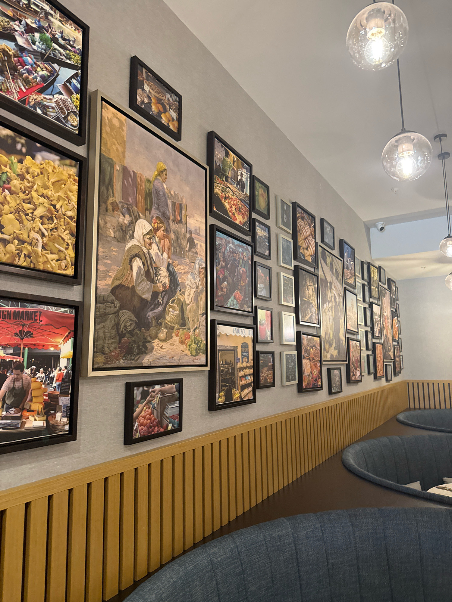 The image shows a wall in a restaurant, likely in Razliv, Bulgaria, decorated with a large collection of framed artwork. The art consists primarily of paintings and photographs, many of which appear to depict market scenes, food, and people in traditional attire. The frames are mostly dark or metallic.
The wall itself is a light gray textured surface. Below the artwork, a wooden paneled wall with vertical slats runs along the length of the scene. In the foreground, curved blue upholstered booth seating is visible, suggesting a dining area.
Hanging from the ceiling are two clear glass pendant lights with visible bulbs, emitting a warm glow. The overall impression is one of a stylish and art-filled dining establishment. No people are clearly depicted engaging in any specific actions within the frame of the image, though some of the artwork itself contains figures. The environment appears to be indoors, well-lit, and clean.