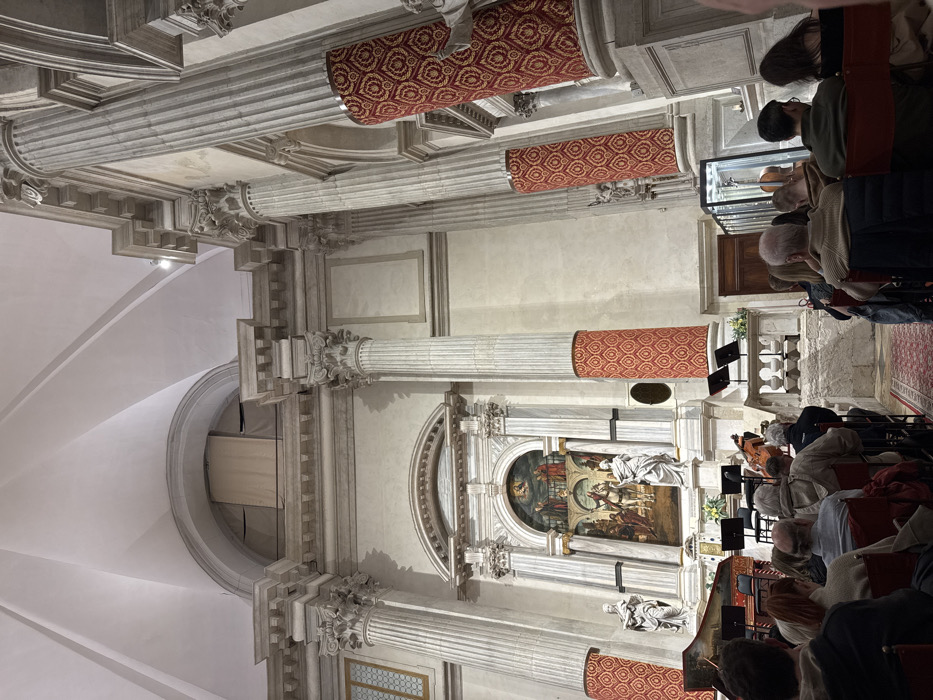The image depicts the interior of the Chiesa di San Polo in Venice, Italy, configured for a musical concert. The perspective is from the audience, looking towards the main altar area, which functions as a stage.
The church features a grand, high-ceilinged Baroque or Renaissance architectural style. The ceiling is white and vaulted, with prominent supporting arches. Fluted columns with ornate capitals, likely Corinthian, line the interior walls. The lower sections of several columns are wrapped in decorative red fabric with a repetitive gold pattern. The walls are light-colored, possibly stone or stucco.
At the center, the main altarpiece is visible. It is an elaborate structure of white marble or stone, featuring smaller columns and an arched niche framing a large painting. This painting is *The Conversion of Saint Paul* by Paolo Veronese, depicting figures on horseback and a heavenly scene. Two white marble statues, identified as Saint Peter on the left and Saint Paul on the right, flank the altarpiece.
In front of the altarpiece, a musical performance setup is present. A dark wooden harpsichord is positioned on the left, and multiple black music stands are arranged. A cellist, largely obscured, is seated and actively playing a cello in the central area. Microphones are visible on stands near the instruments.
A seated audience occupies the foreground and midground, facing the performance. Individuals are primarily seen from behind, occupying rows of red chairs. The ambient lighting illuminates the architectural details and the performance area.
