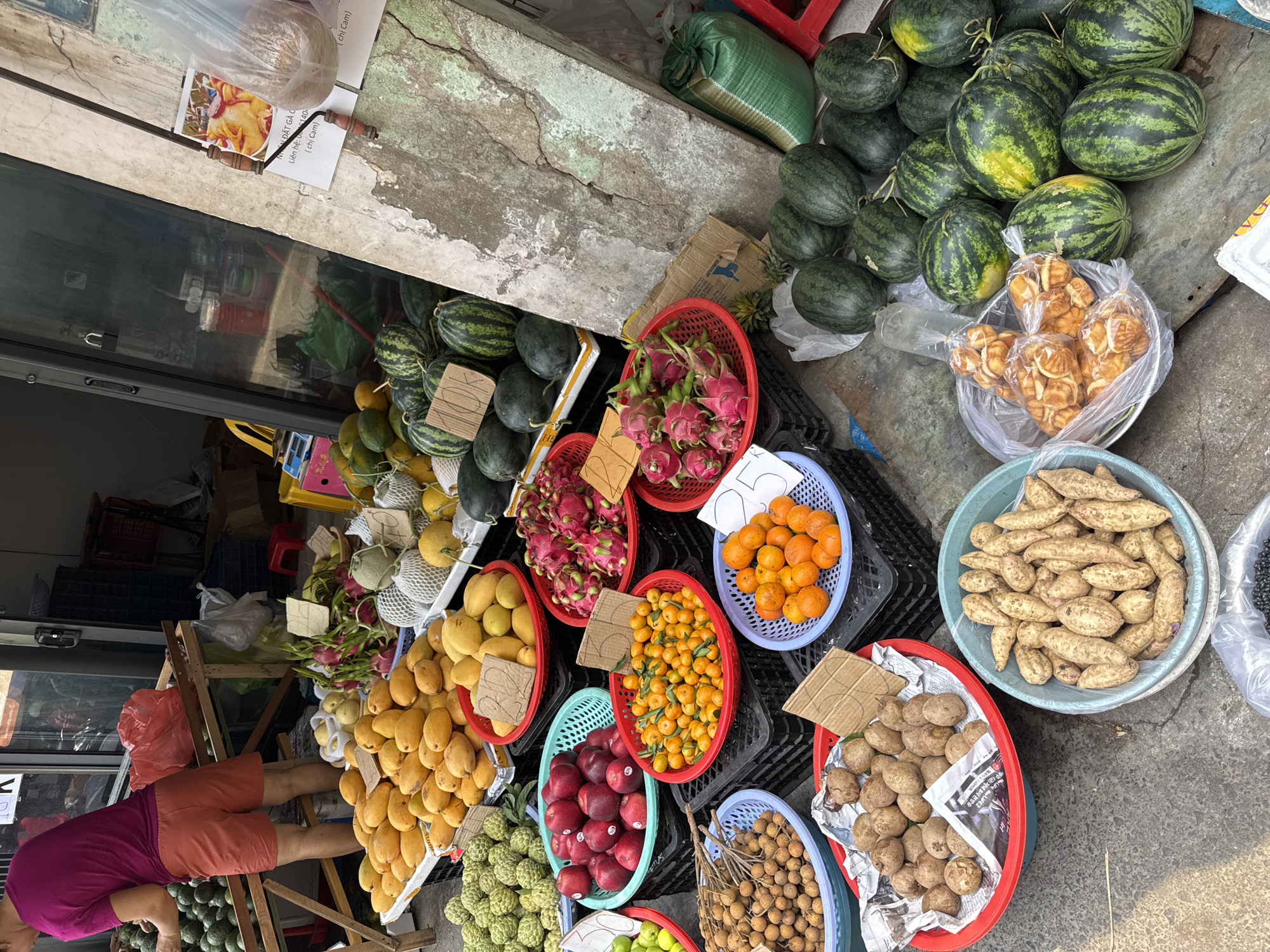 The image depicts a bustling fruit and vegetable market scene, likely in Đà Nẵng, Vietnam. A variety of fresh produce is displayed in colorful plastic baskets. The assortment includes watermelons, mangoes, dragon fruit, oranges, and various types of smaller fruits, such as longans. Sweet potatoes and other root vegetables are also present. Some items are accompanied by handwritten price tags. A vendor, visible only from the knees down, stands near the produce. A sign with Vietnamese text and a phone number is partially visible, suggesting a local business or service. The market setting appears to be outdoors, with the products arranged on the ground and against a storefront.
