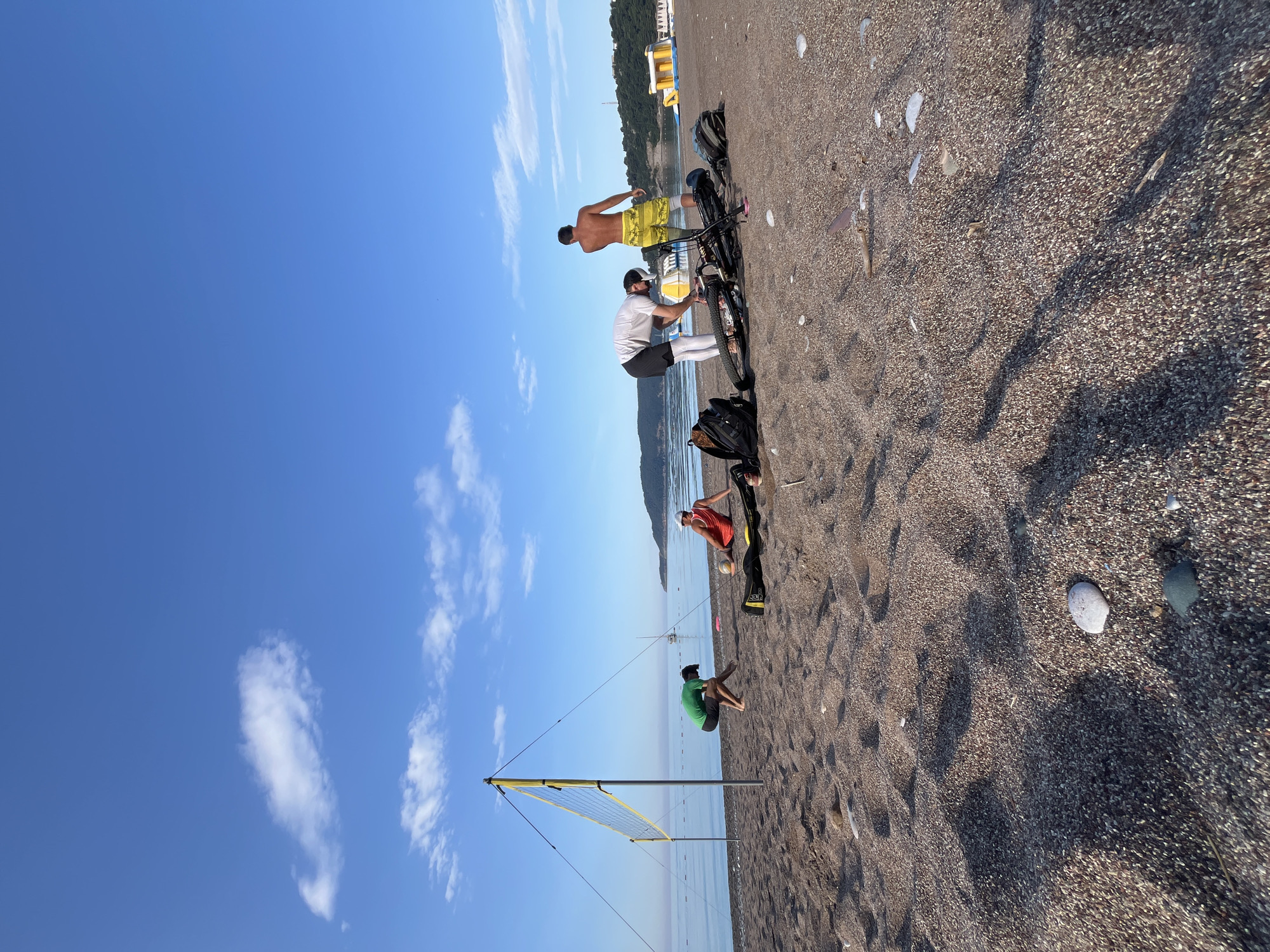 The image captures a beach scene in Bečići, Montenegro. In the foreground, a volleyball net is erected on the sand. A person in green clothing is seated near the net. Further back, two people are standing and playing with a volleyball, with another individual present. A bicycle is parked on the beach nearby. The background includes the Adriatic Sea and the sky. The beach is composed of fine dark sand and pebbles.