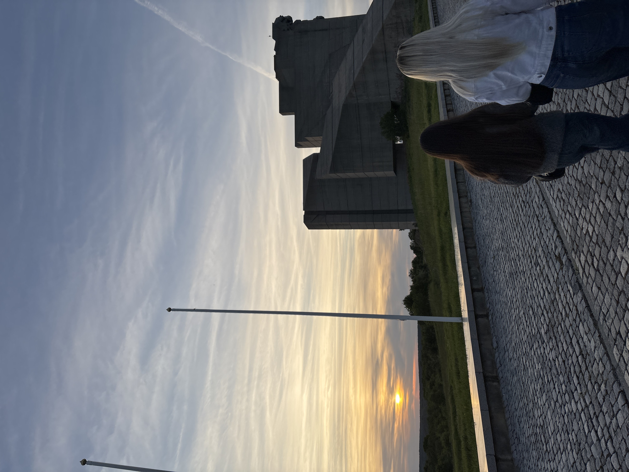 The image shows the Monument to the Buzludzha Congress in Bulgaria at sunset. The concrete structure stands in the background with two individuals facing away from the camera to the right of the monument. The foreground features a cobblestone paved area and flagpoles. The sun is setting on the horizon, casting an orange glow over the landscape.