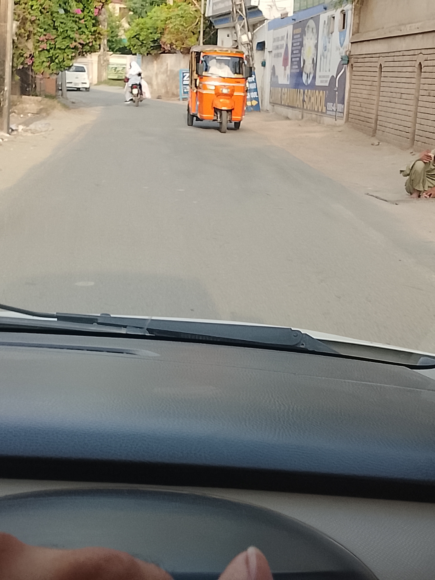 The image, taken from inside a vehicle (dashboard and windshield wipers visible), shows a street scene in Gulgasht Colony, Pakistan. An orange auto-rickshaw is traveling in the opposite direction on the street. In the distance, a person on a motorcycle is seen. To the right, near a brick wall, a person is sitting on the ground. On the wall, a sign for Grammar School is visible. Other vehicles are visible in the background.