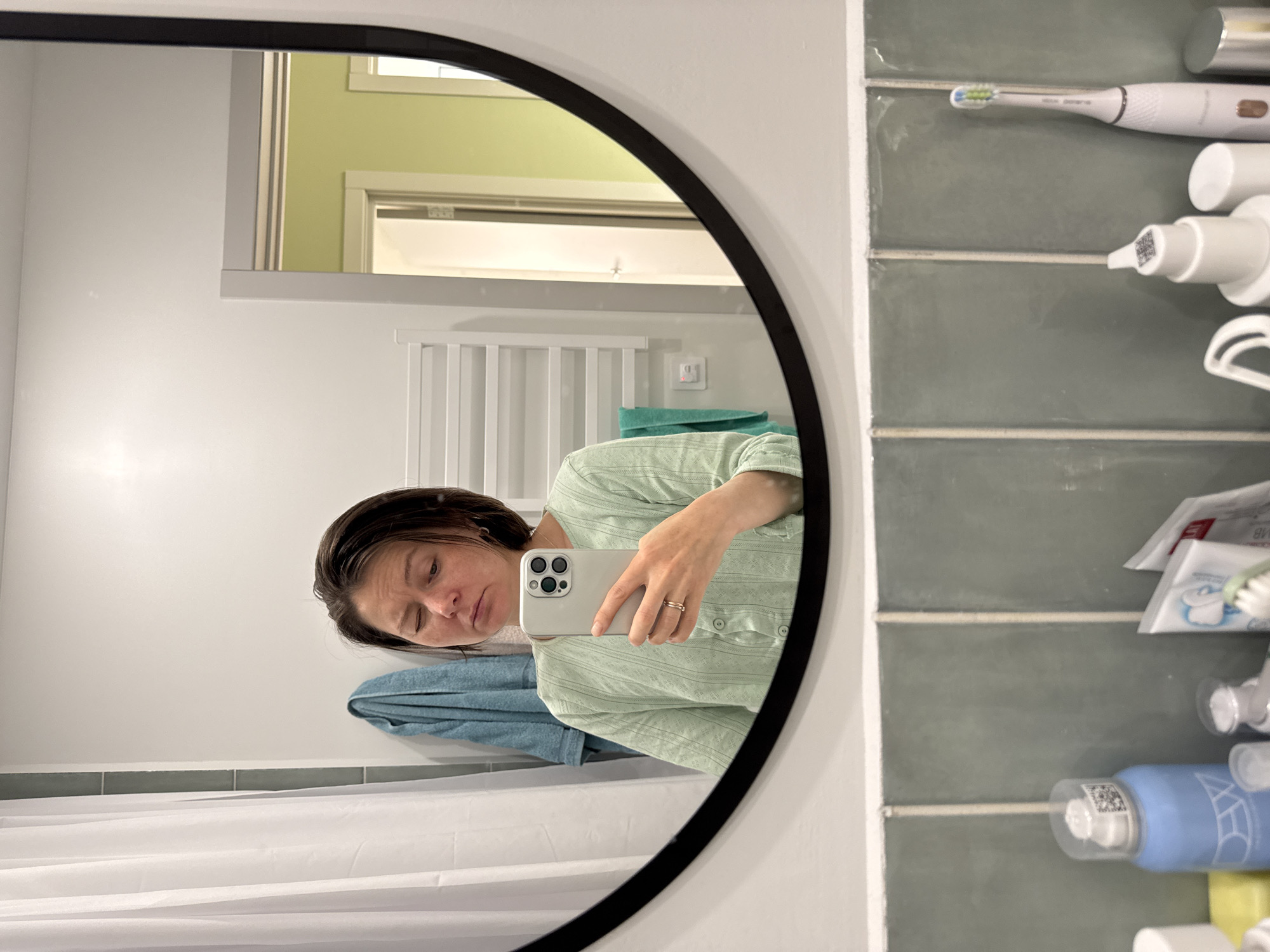 A selfie taken in a bathroom, reflected in a round mirror with a black frame. A woman wearing a light green, button-down shirt is holding a smartphone to take the picture. A white shower curtain hangs in the lower left corner. A blue towel is visible behind the woman. Behind her in the reflection, there is a white, heated towel rack. On the right side of the frame, there is a wall with large, light green tiles, holding an electric toothbrush and various toiletries. The location is presumably Люк, Russia.