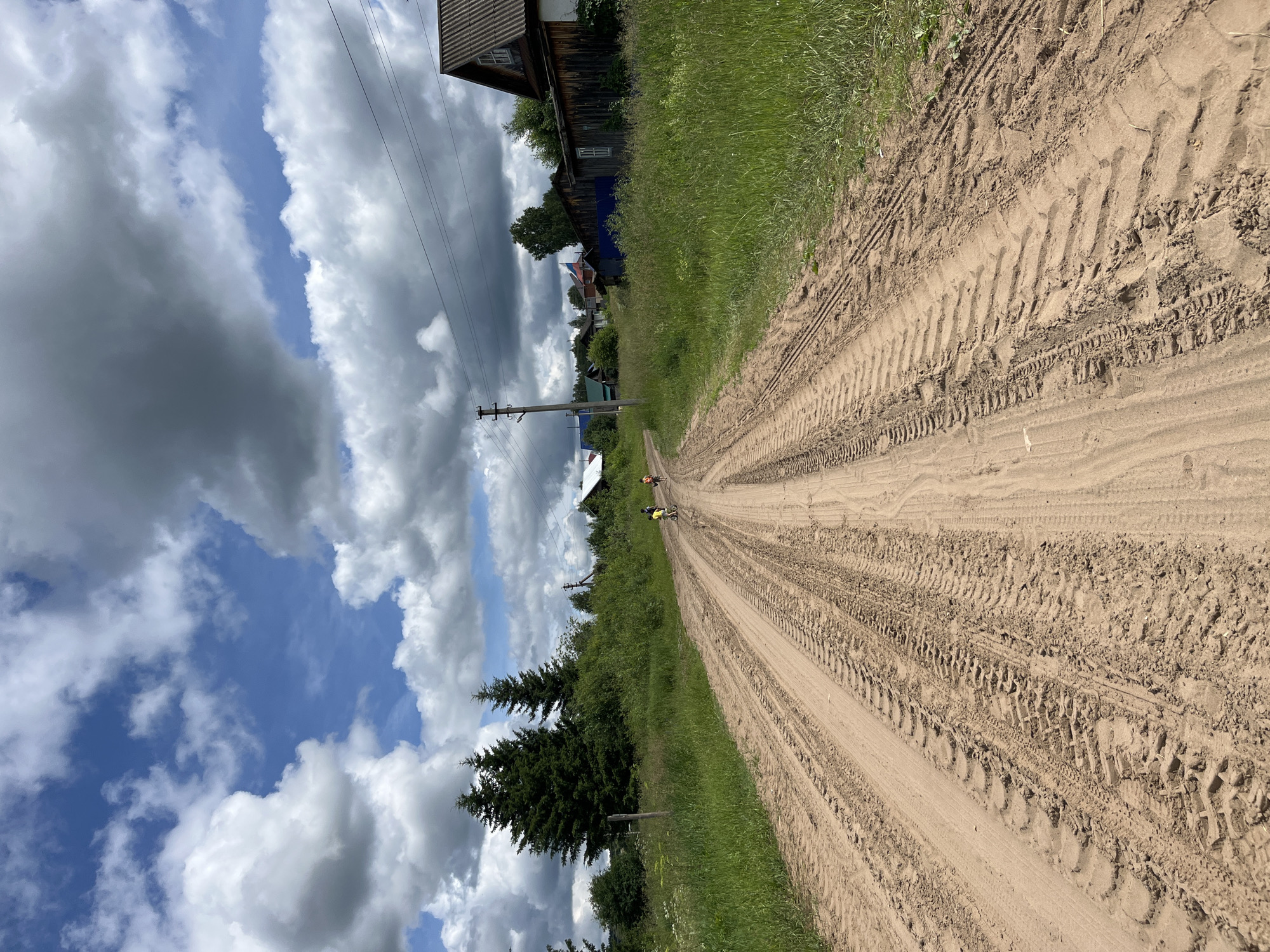 A dirt road, marked with tire tracks, stretches into the distance, bordered by lush green grass and foliage. In the middle ground, two figures on bicycles are riding away from the camera. A few scattered houses and trees line the right side of the road. The sky is a mix of blue with dramatic, fluffy white and grey clouds. The scene appears to be in a rural area, possibly the outskirts of Уральское, Russia, given the presence of wooden houses and a dirt road characteristic of such locations. The lighting suggests it is daytime, with the sun intermittently breaking through the clouds.