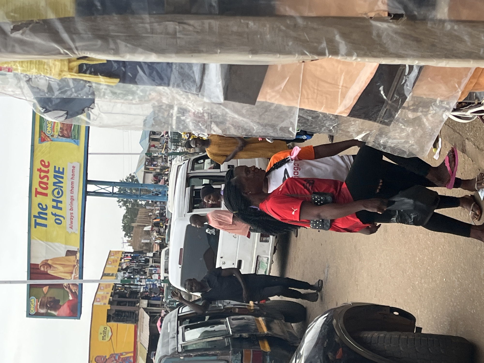 A street scene in Bukuru, Nigeria, features several individuals amidst vehicles and market stalls under a cloudy sky. In the foreground, a person in a red shirt and black leggings is visible from the waist down, carrying a black plastic bag and wearing flip-flops. To their left, a black SUV is partially visible.
Behind these individuals, a dark colored van is parked on the left, with a man in a black t-shirt and pants waving. Next to the van, a white minivan is positioned, with a man in a pink t-shirt standing nearby and another man in a yellow traditional tunic standing to the right of the minivan. In the background, market stalls are set up, displaying various goods. A large billboard advertising "The Taste of HOME" by ONGA is prominently displayed above the market. The ground is paved with dirt and asphalt.