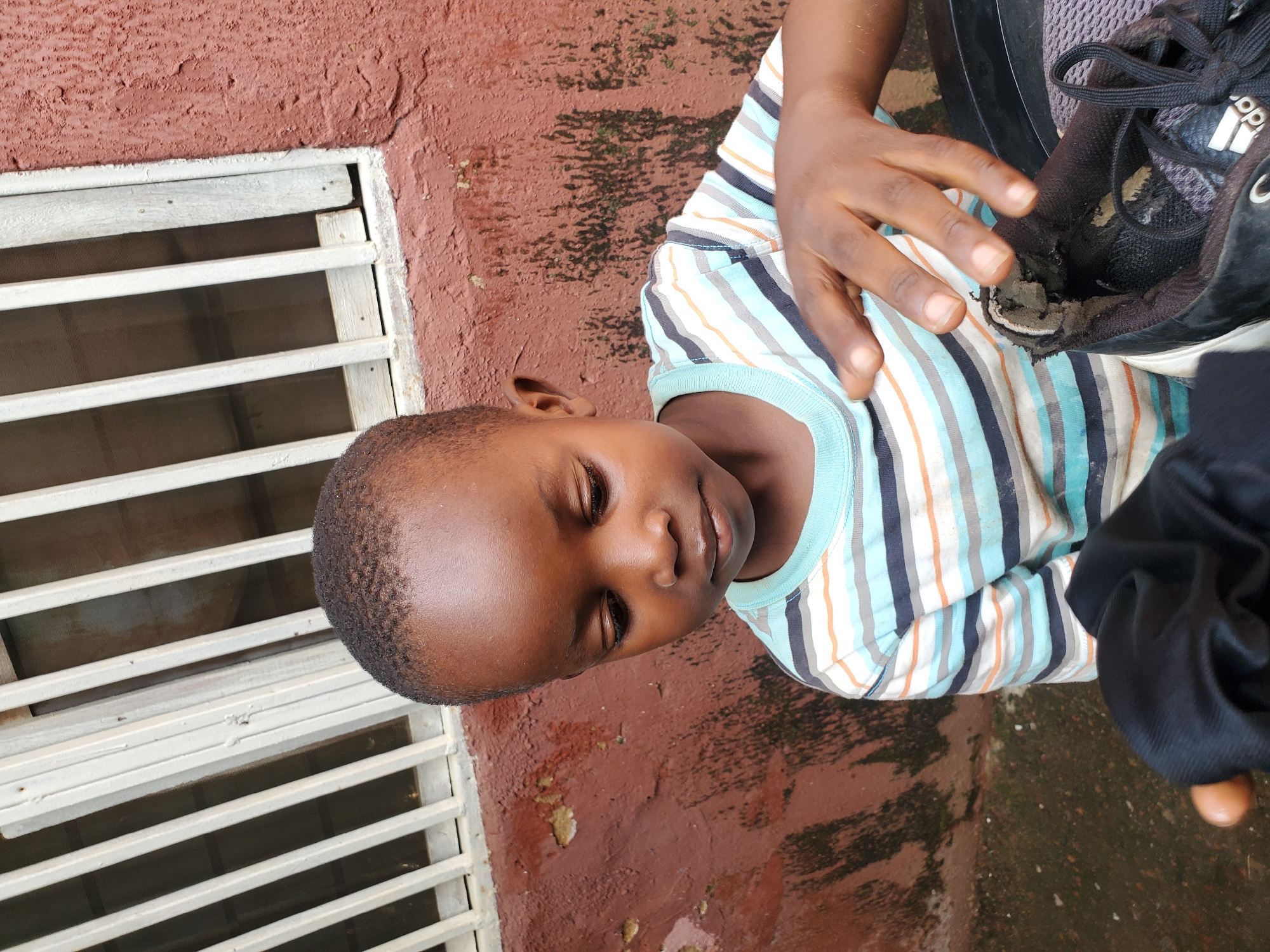 The image shows a young boy in Jos, Nigeria. He is lying down on a reddish-brown, textured surface that appears to be the ground or a wall. To his left, there is a white metal grate or window covering. The boy is wearing a striped t-shirt with blue, white, orange, and black stripes. His eyes are closed, and he appears to be resting or sleeping. His right hand is resting on a black shoe, which looks worn and damaged. The lighting suggests it might be daytime, possibly overcast or in the shade, as there are no strong shadows. The overall scene gives a sense of everyday life. No visible text is present in the image.