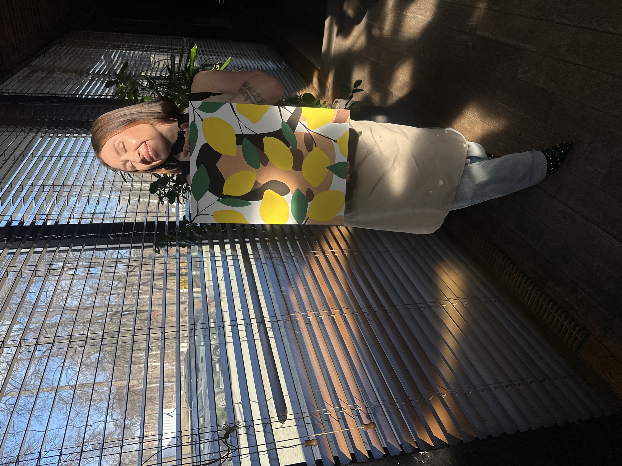 A woman is shown standing in front of a window with blinds, holding a painting of lemons and foliage. She is wearing jeans, a light-colored apron with paint splatters, and black shoes with studs. She is facing slightly towards the left of the frame and sticking her tongue out playfully. The painting she holds covers part of her torso. Behind her and to the side, plants are visible. The background shows a cityscape through the blinds. The location could be Izhevsk, Russia, but there is no direct confirmation in the image. The light suggests it is daytime. The floor is made of dark wood.