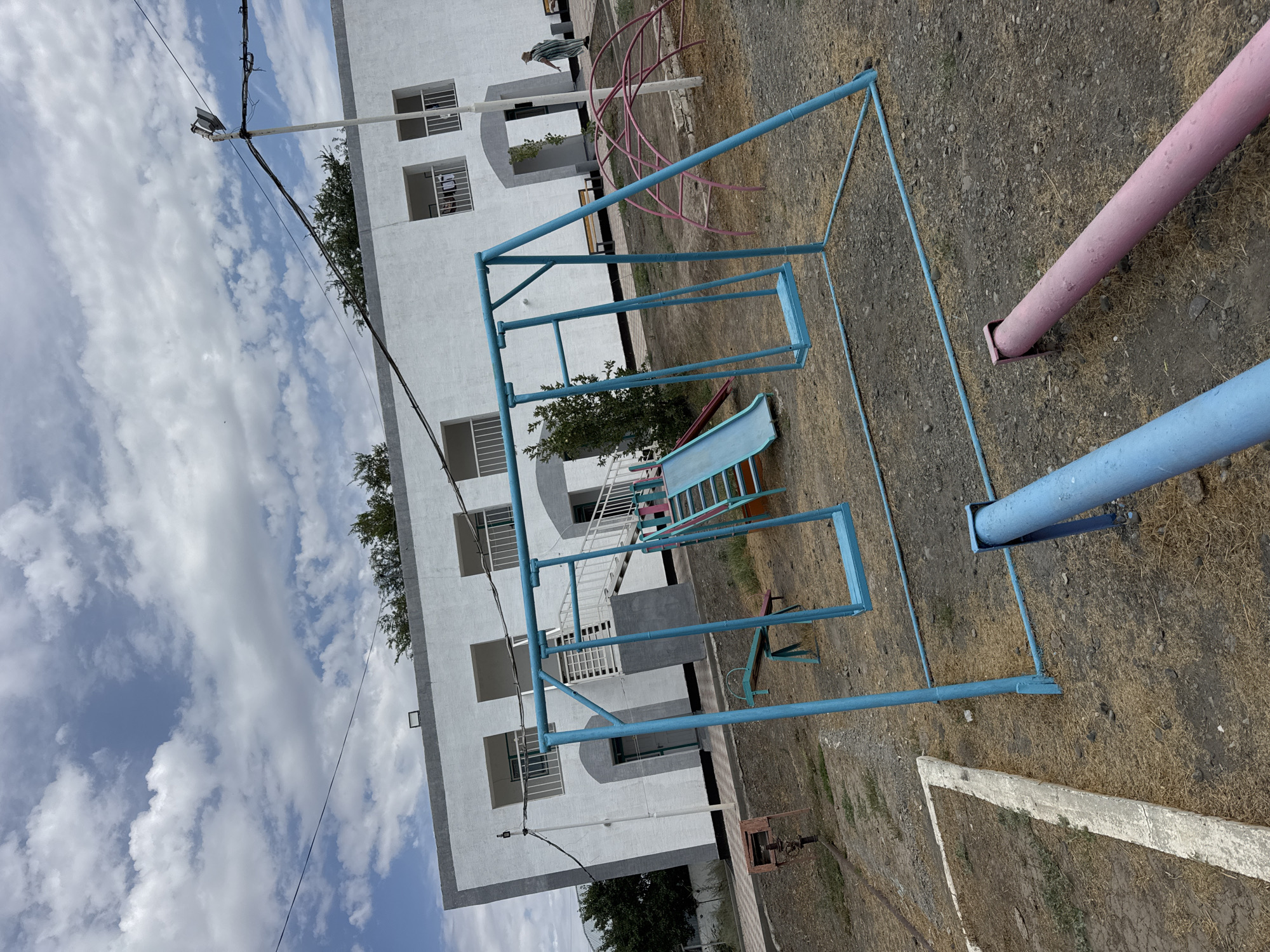 The image depicts an outdoor playground area in front of a white, two-story building. The building has multiple square windows with railings and a central entrance with a staircase. The playground features a blue metal swing set with two swings, a blue and green slide, and a pink arched climbing structure. The ground is covered with dry grass and gravel. A person in a striped garment is walking in the background near the building entrance. Power lines are visible overhead. The scene appears to be located in Zharbulak, Kazakhstan, as indicated by the context. The overall atmosphere is one of a slightly neglected but functional public space.