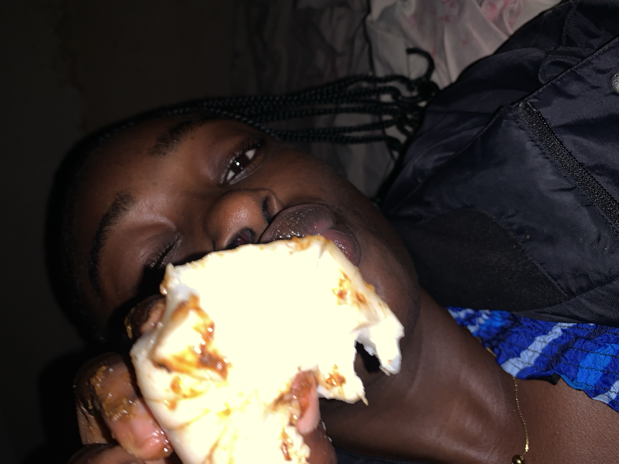 A close-up shot shows a person of African descent eating. The person is holding a piece of food up to their mouth, with their tongue extended to lick the food. The food appears to be white and covered in a reddish-brown sauce. The person is wearing a dark jacket over a blue and white patterned top, and a thin gold chain. Their hair is braided and visible on the right side of the frame. The background is dark and appears to be a bed or couch, suggesting an indoor setting. There are no identifiable landmarks or contextual clues to pinpoint a specific location in Rafin Par, Nigeria.
