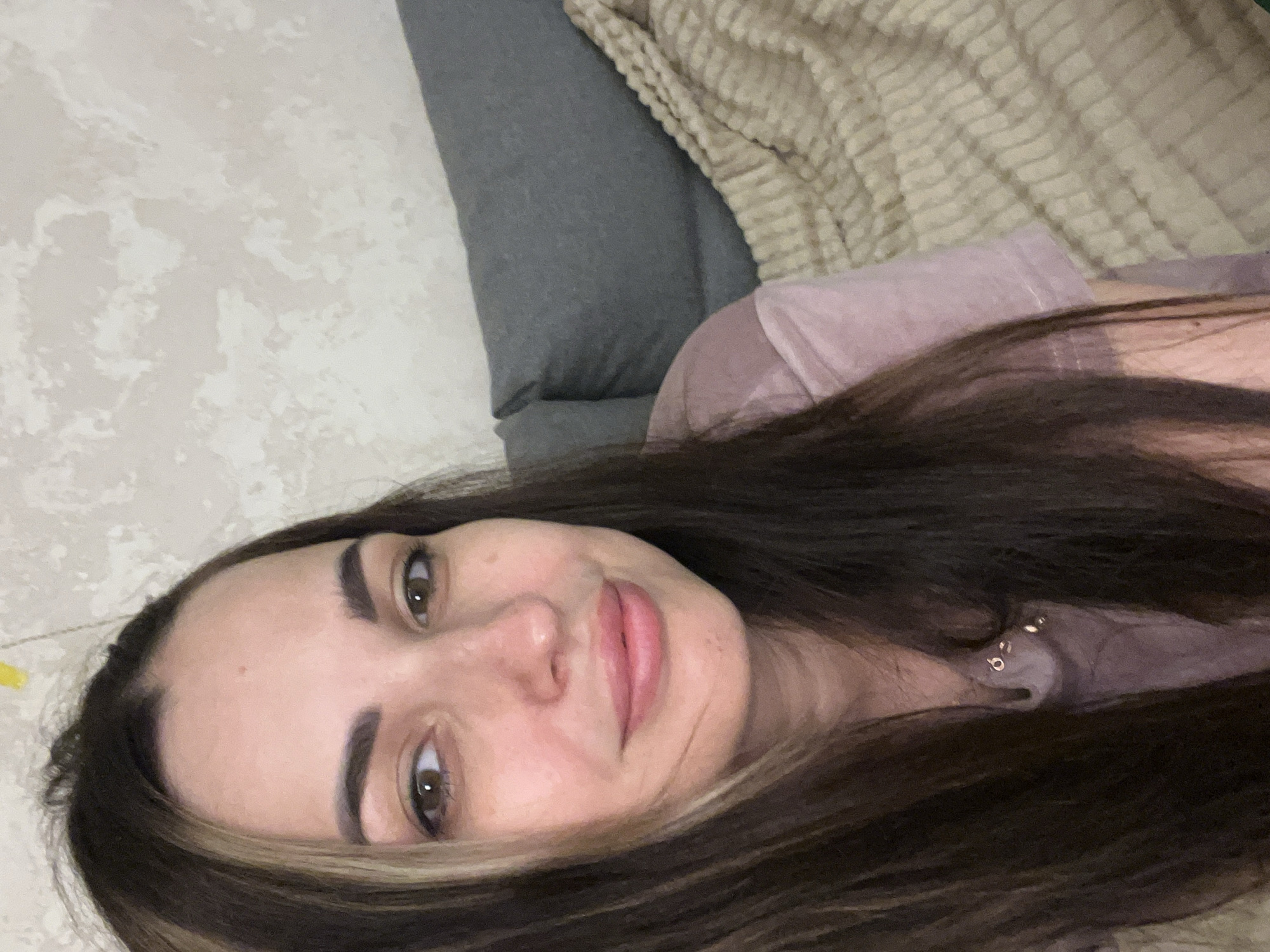 A medium close-up selfie of a woman with long, dark brown hair and prominent highlights is captured. She has fair skin, light brown eyes, and is wearing a pink t-shirt. She is looking directly at the camera with a slight smile. The background consists of a textured, light-colored wall and a grey sofa with a beige knitted blanket draped over it. The overall setting appears to be indoors, possibly a living room.