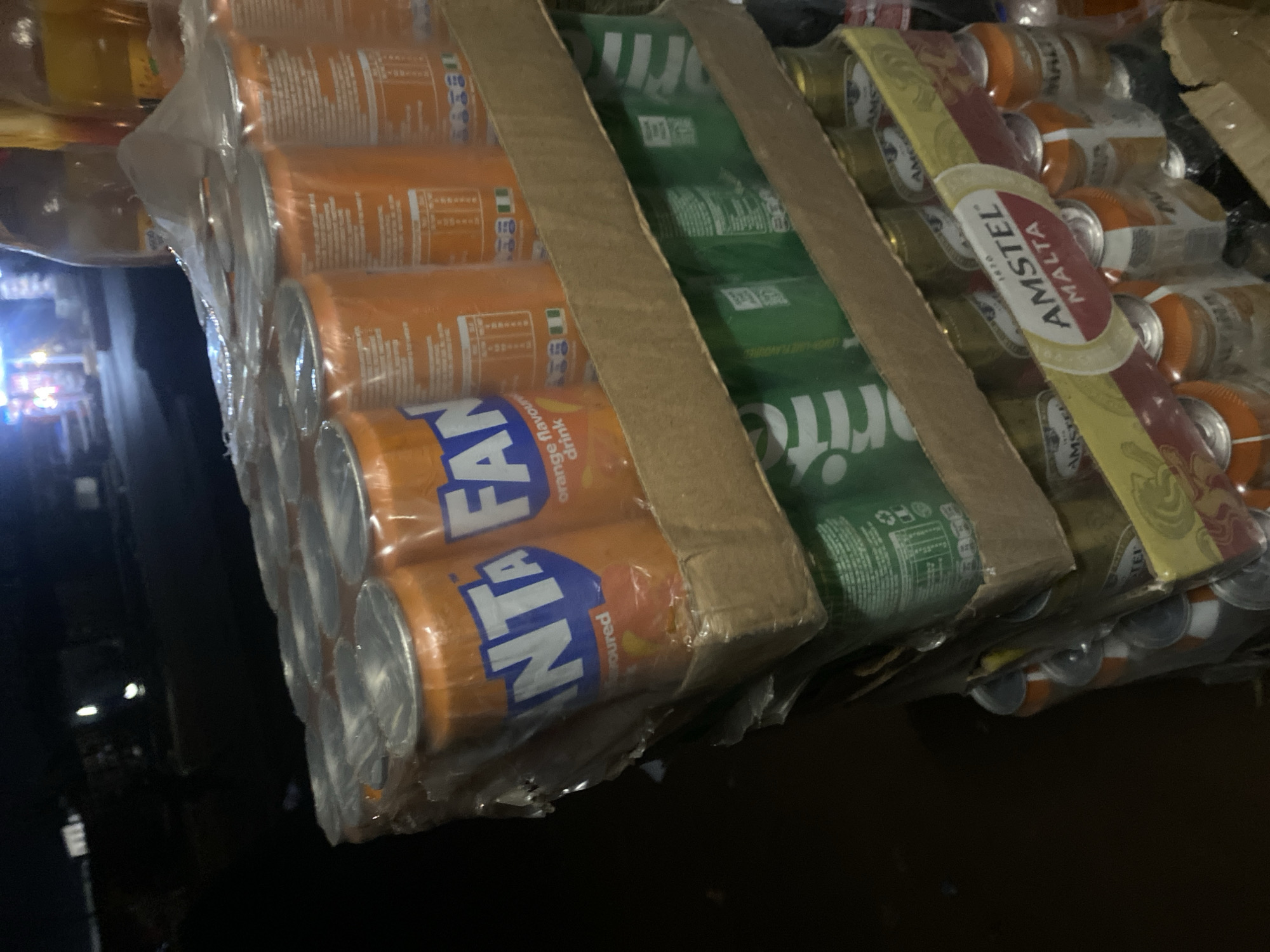 The image displays a vertical stack of commercially packaged beverages, presented on cardboard support structures. The lighting suggests a dimly lit environment, possibly night or an indoor commercial space, with blurred artificial lights visible in the distant background.
The top layer consists of multiple shrink-wrapped multi-packs of orange-flavored Fanta cans. These cans are orange with prominent blue "FANTA" branding. On the reverse side of several cans, nutritional information panels are visible, along with a small green, white, and green flag icon, identifying the product's market as Nigeria.
Immediately below the Fanta layer, a stack of green-colored beverage cans, consistent with Sprite branding, is visible, also secured in clear plastic multi-packs. The white "Sprite" logo is partially discernible on these cans.
The lowest visible layer comprises golden-colored Amstel Malta cans, similarly arranged in multi-packs. The "AMSTEL MALTA" branding is clearly displayed on red and yellow labels. Beneath this layer, several individual bottles with orange caps are partially visible.
No individuals are present within the frame. The scene is consistent with a retail or vendor display of popular beverages, commonly found in commercial settings within Nigeria, as indicated by the national flag on the Fanta cans. While the provided context specifies the location as Ikorodu, Nigeria, the image itself does not contain any unique landmarks or specific identifying features to confirm the precise location within the city.