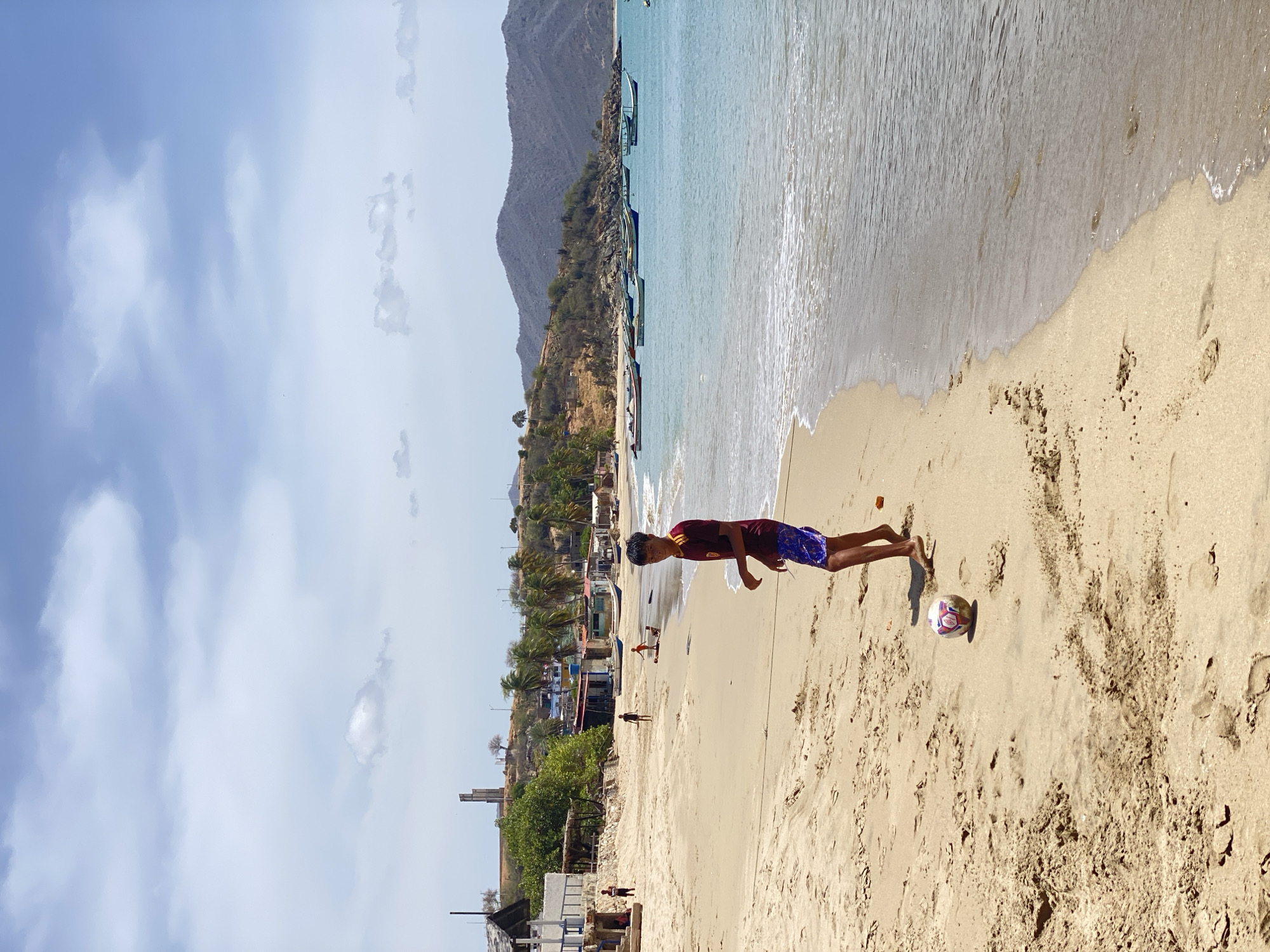 The image captures a vibrant beach scene under a bright, partly cloudy sky. The main subject is a young male, positioned prominently in the mid-foreground on the sandy beach, facing towards the viewer. He wears a dark red t-shirt and blue patterned swim shorts, standing barefoot with a colorful soccer ball resting near his feet on the sand.
The broad sandy beach stretches towards the right, meeting the calm, clear turquoise waters of the ocean, where gentle waves lap the shore. Along the coastline in the mid-ground, several traditional-looking fishing boats, often called *peñeros*, with colorful trim, are either pulled onto the sand or gently bobbing in the shallow water.
Beyond the immediate beach, a rugged, arid-looking hillside rises, covered with sparse vegetation, including some palm trees. Scattered small buildings and structures are visible along the base of the hill, suggesting a coastal settlement or village. In the distant background, large, hazy mountains complete the picturesque coastal landscape.
Several other smaller figures are sparsely distributed on the beach in the mid-ground, indicating other people enjoying the area, though their specific activities are not clearly discernible. The scene is bathed in bright daylight, suggesting a sunny day, likely late morning or early afternoon, with minimal shadows. No visible text is present in the image. The setting conveys a serene and recreational atmosphere at a coastal location, identified as Juan Griego, Venezuela.