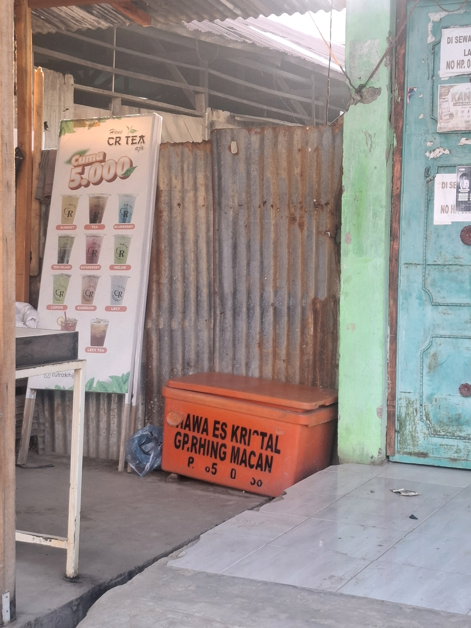 The image depicts an outdoor scene in front of a building, possibly a shop, in Mesjid Tuha, Indonesia. The foreground shows a cracked concrete floor with light-colored tiles. To the left, a white metal frame with wooden supports suggests a stand or table.
Dominating the center is a large, orange insulated box, likely a cooler, with black lettering that reads "MAWA ES KRISTAL GP.RHING MACAN P. 05 0 30". To the left of the cooler, a blue plastic bag is visible on the ground.
Behind the cooler and to the left, a tall, white sign advertising drinks is propped up against a corrugated metal wall. The sign displays various drinks with names like "SUNIST", "TEA", "BLUEBERRY", "TEH HIJAU", "ROSEBERRY", "MELON", "LEMON", "RAINBOW", and "LECY". The prominent text on the sign reads "Cuma 5.000" and "CR TEA".
The corrugated metal wall extends across the midground, showing signs of rust and age. To the right of the corrugated wall is a brightly painted green pillar, and further to the right is a teal-colored door with ornate carvings and dark metal accents. Advertisements or notices are affixed to the door and the green pillar. The upper portion of the image reveals a weathered metal roof supported by wooden beams, suggesting a simple, utilitarian structure. The lighting indicates it is daytime, with sunlight casting shadows and creating a slightly hazy atmosphere.