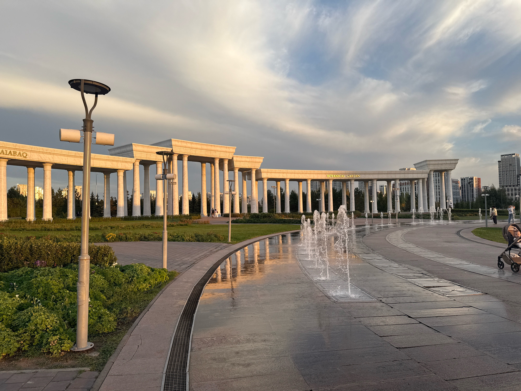 This is an outdoor, eye-level, wide-angle shot of the Botanical Garden in Astana, Kazakhstan, during the late afternoon or early evening. The sky is a mix of white and gray clouds with patches of blue, illuminated by the setting sun. In the background, a long, curved colonnade with white marble columns extends across the frame. Above the colonnade, text is visible, with "BOTANICAL GARDEN" clearly readable on the right side, and "MALABAY" on the left. Behind the colonnade, modern high-rise buildings are visible.
In the foreground, a wet, paved walkway with a curving pattern of large, dark gray tiles runs from the right to the left. A series of water fountains are active in the middle of the walkway, shooting streams of water into the air. The wet surface of the walkway reflects the light and the colonnade. To the left of the walkway, a strip of green grass and manicured bushes and flowers is visible. Two tall, modern streetlights with circular tops stand on the left side of the frame, one closer to the viewer and one further back.
On the right side of the image, a paved path curves away from the viewer, and a person is pushing a stroller with a child in it. Further back on the right, another person stands near the colonnade. On the left side of the colonnade, in the distance, two figures can be seen walking. The overall impression is one of a well-maintained public space with architectural grandeur and a serene atmosphere.