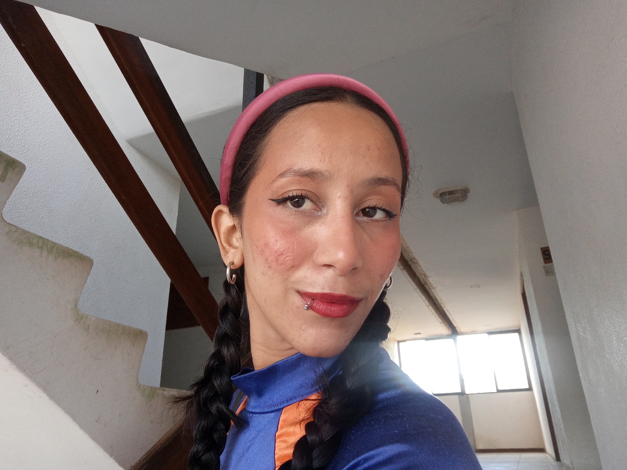 A medium, close-up shot shows a person standing indoors. The subject appears to be female, has dark hair styled in two braids, a lip piercing, and wears a pink headband, hoop earrings, and blue and orange clothing. She stands on a landing in front of a white wall, with brown wood and white stairs visible in the left background. The landing leads to a hallway with a window in the right background. The location appears to be a residential or institutional building in Porlamar, Venezuela.