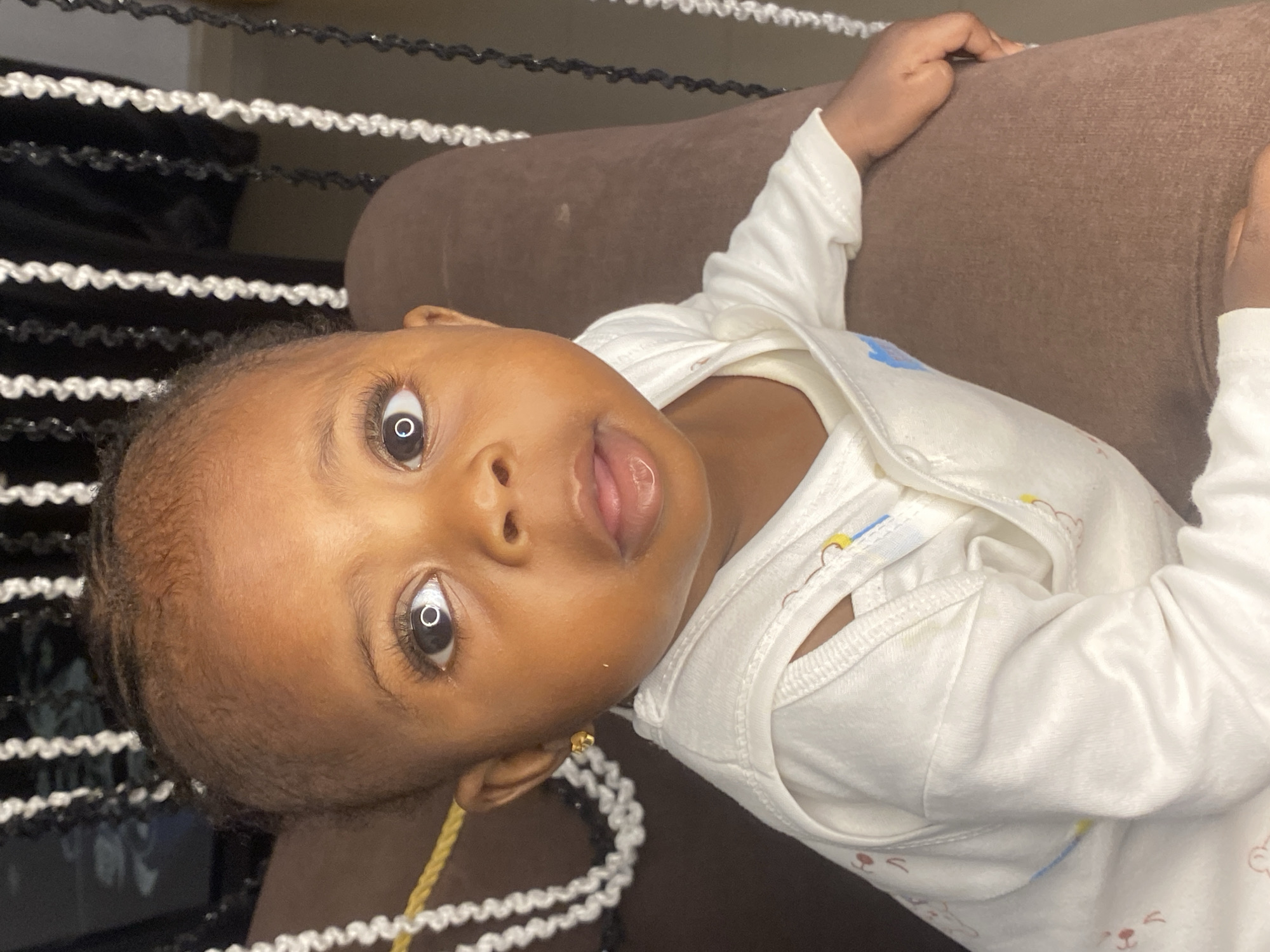 A close-up, head-and-shoulders shot shows a young Black child wearing a white onesie adorned with faint illustrations of animals. The child has dark hair styled in braids, brown eyes with long lashes, and a small gold earring in their left ear. Their mouth is slightly open, with a hint of moisture on the lower lip. The background consists of vertical strands of white and black decorative beads, hanging against a dark surface. A brown, textured surface, likely a piece of furniture, occupies the right side of the frame, against which the child is resting. The scene appears to be indoors, possibly in a residential setting within Abakaliki, Nigeria.