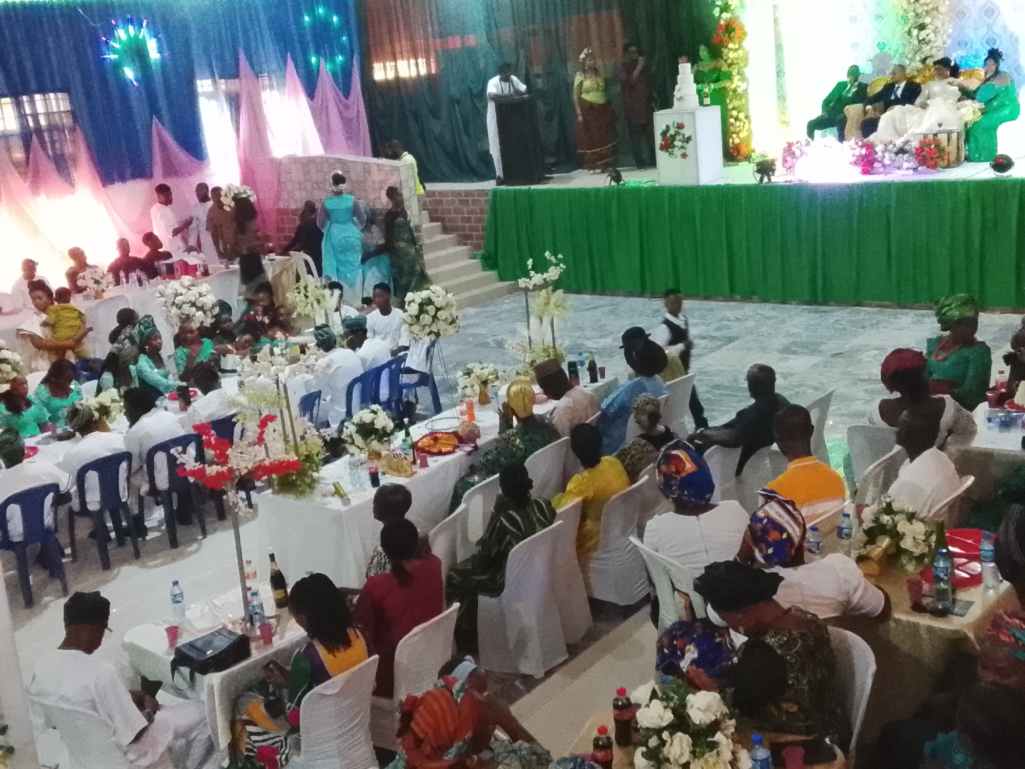 The image captures a scene at a wedding reception in Jos, Nigeria. The reception hall is decorated with white tablecloths, floral centerpieces, and white chairs. A raised stage area is visible in the background, where the wedding party sits, and a speaker stands at a podium. Several guests are seated at tables throughout the hall, engaging in conversation. Some are turned towards the stage, indicating they are listening to the speaker. The hall is decorated with draping fabrics in various colors.
