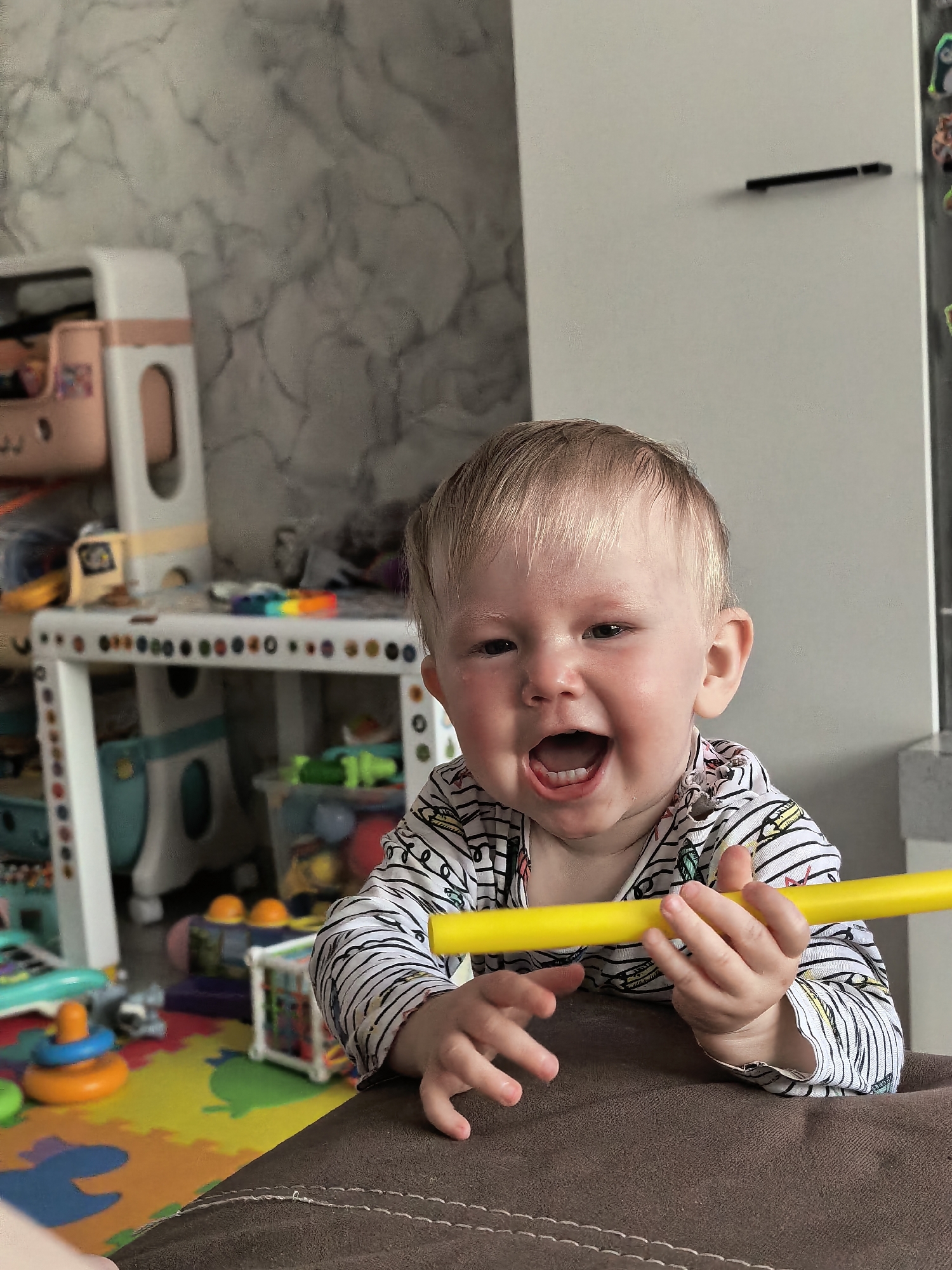 A fair-skinned baby with blonde hair, wearing a striped shirt with colorful patterns, is holding a yellow rod and smiling with their mouth open. The baby is leaning on a brown surface with visible stitching. In the background, there's a play area with a colorful puzzle mat, toys, and a small white table. A gray marble-patterned wall is visible behind the table. The scene is indoors, with a white cabinet or wall segment in the background. There's no specific indicator of location in Sterlitamak, Russia.