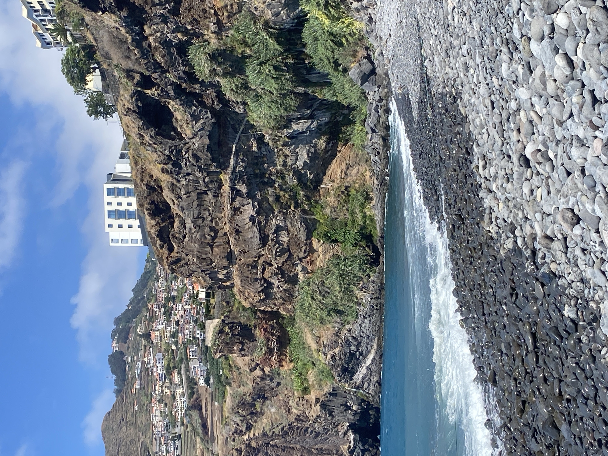 The image captures a view of a pebbled beach in Câmara de Lobos, Portugal, with the ocean in the foreground and a steep, rocky cliff face behind it. The water is a vibrant blue, with small waves breaking on the shore. The beach is covered in smooth, dark pebbles, with some lighter-colored ones scattered throughout.
The cliff face is rugged and layered, with patches of dry vegetation and scrub growing in its crevices. At the top of the cliff, houses with red-tiled roofs are clustered on the hillside, characteristic of coastal villages. Several modern buildings, including a white structure with a grid of windows and a taller, multi-story building with balconies, are visible on the upper slopes. The sky is blue with scattered white clouds. The scene is natural and offers a panoramic view of the coastline and the settlement. There are no people or animals visible in the frame.