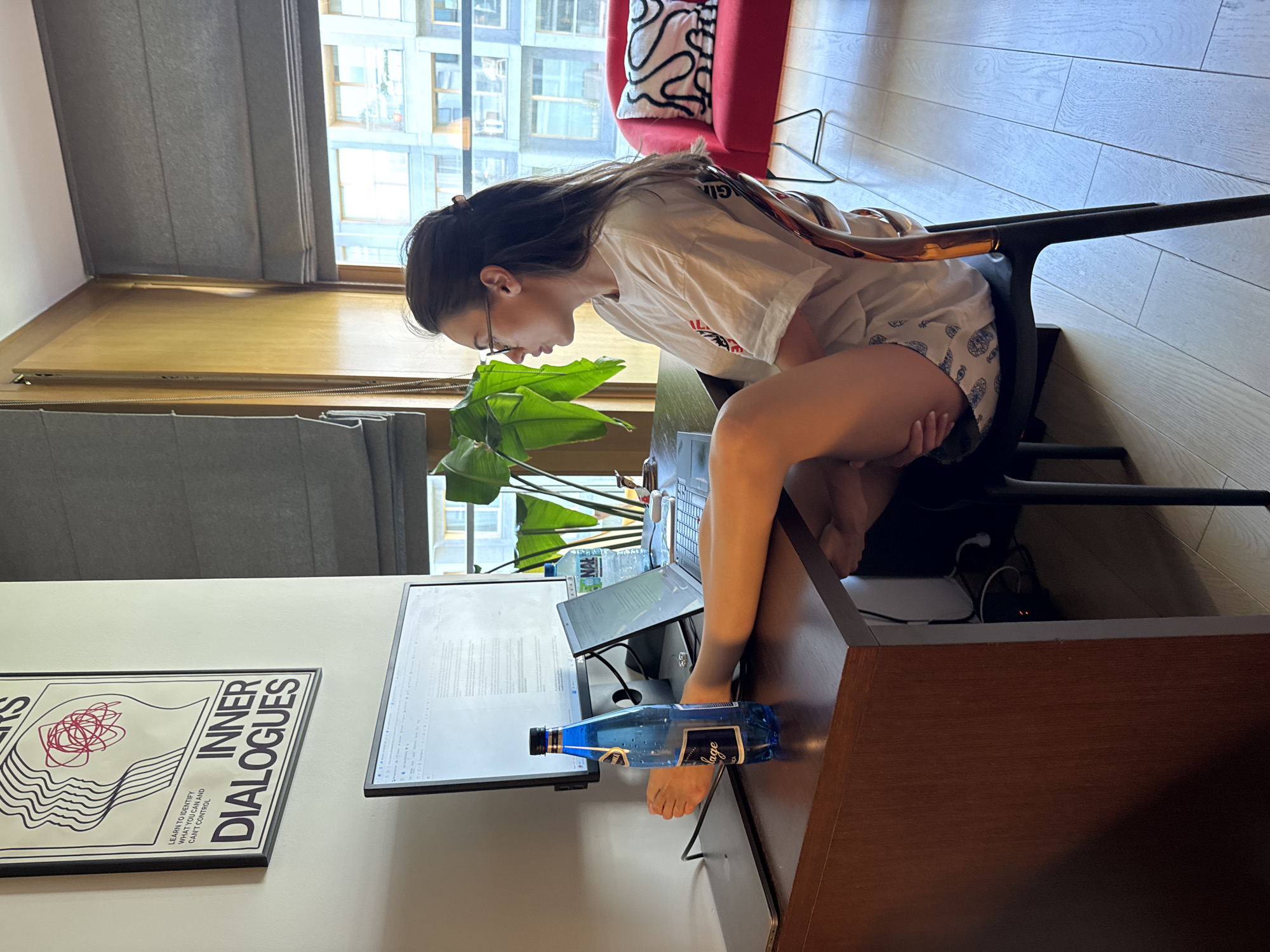 A young woman with long, dark hair sits at a desk in an indoor setting, likely an apartment. She is wearing glasses, a white t-shirt, and patterned shorts. The woman is using a laptop computer, which is positioned on a desk in front of a large computer monitor displaying text. Next to the monitor is a poster with the words "INNER DIALOGUES" printed on it. A bottle of water is placed next to the monitor. A green potted plant sits behind the computer. In the background, a window reveals a cityscape, and a red chair with a patterned pillow is partially visible. The floor is made of light-colored wood.