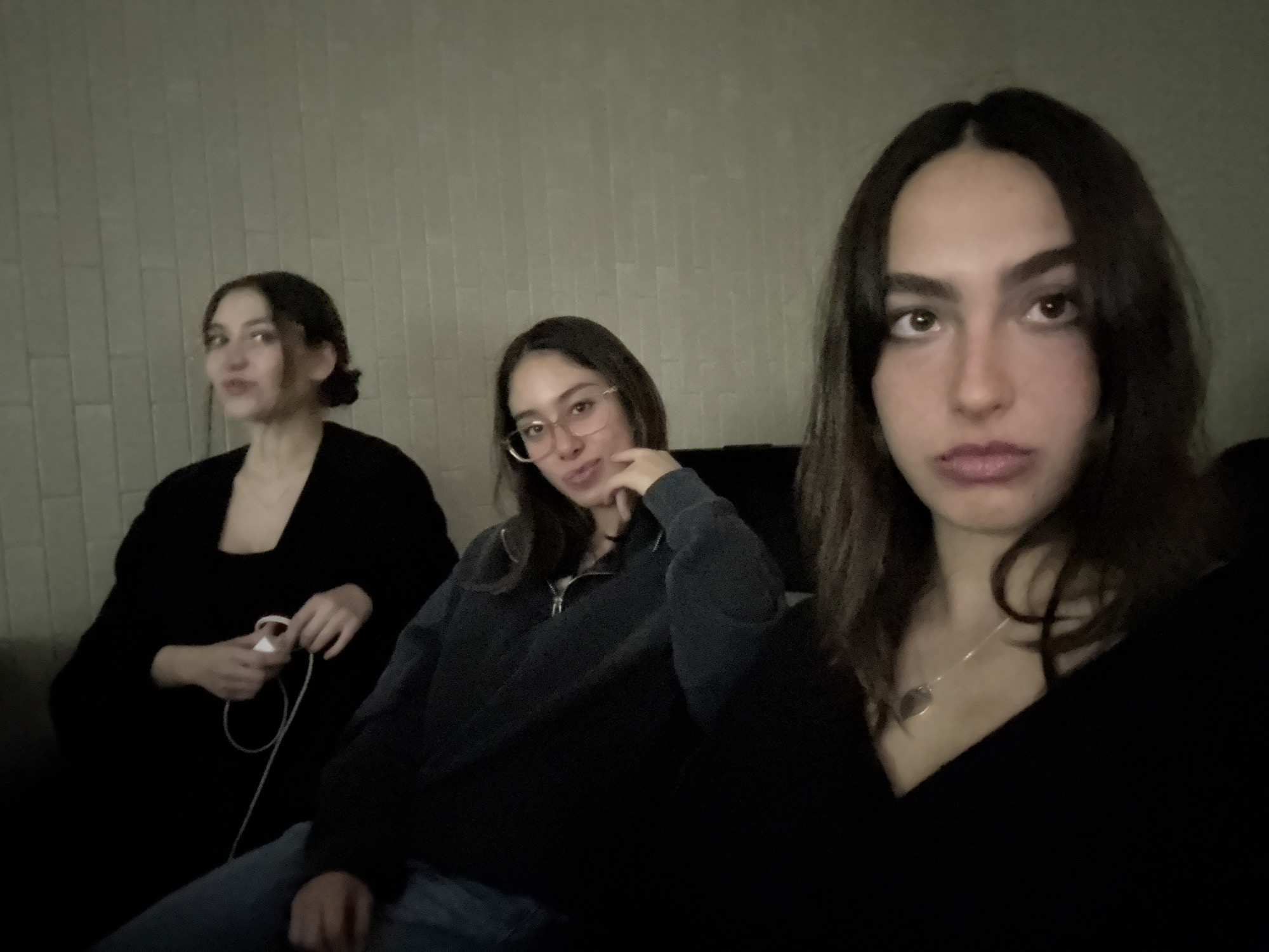 Three young women are seated indoors against a light-colored, textured wall that resembles brick or tile. The woman on the left has her hair up and is wearing a black top, holding a white object with a cord. The woman in the middle wears glasses, a dark zip-up jacket, and jeans, resting her chin on her hand. The woman on the right has long dark hair, is wearing a black top and a necklace with a pendant. The overall scene has low lighting. The location is likely a residential or entertainment space indoors, though there is no evidence indicating it is located in Huixquilucan de Degollado, Mexico.