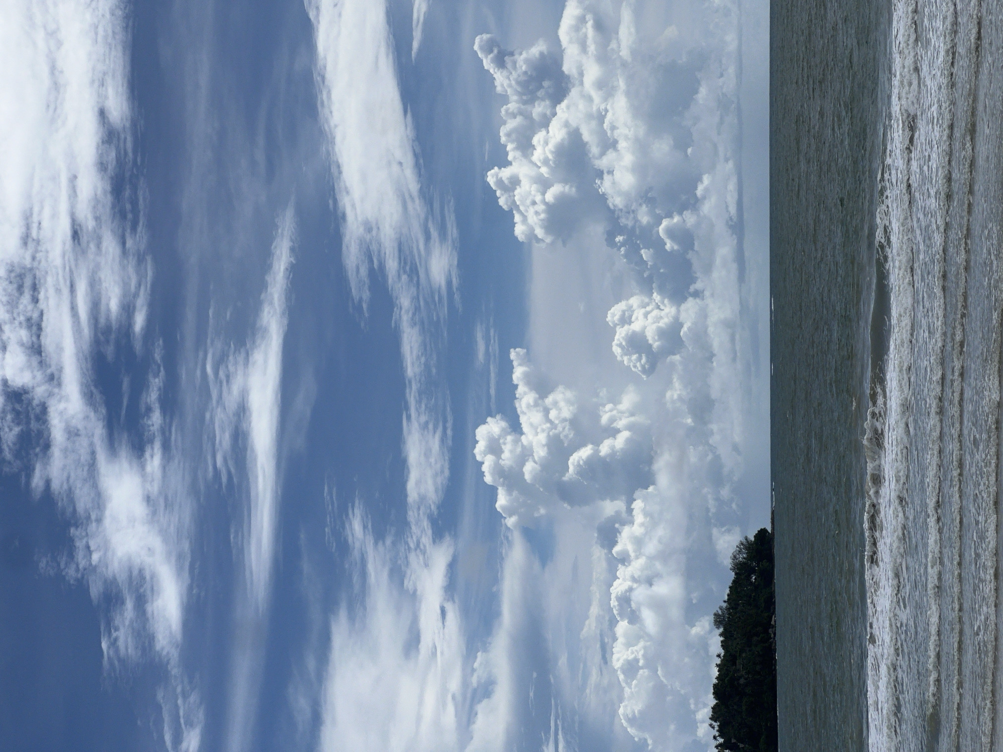 A full shot, eye-level, outdoor, vertical, medium-distance photograph shows a tropical island in the ocean under a bright, cloudy sky. The island is on the far left of the frame and is covered with dense, dark green trees, with some exposed rock formations visible. The ocean is a dark blue-grey color with small white-capped waves rolling towards the shore, which is not visible. The sky is a bright blue with large, puffy white clouds, some of which are tinged with grey, indicating potential rain. The horizon line is clear, dividing the ocean and sky. The scene appears to be in a tropical region, given the lush vegetation on the island. The overall impression is one of a serene, natural landscape.