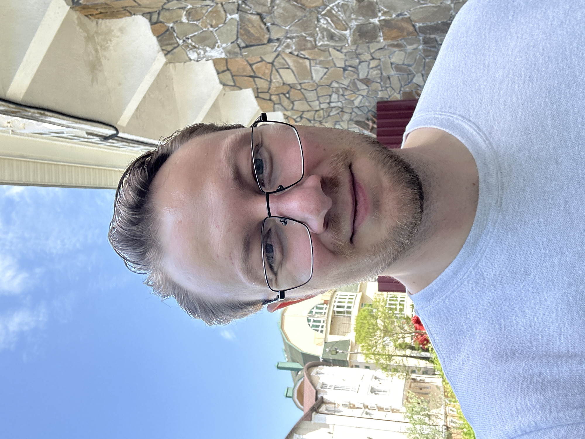 A close-up, selfie-style photograph shows a man wearing glasses and a white t-shirt, seemingly outdoors. Behind him, there's a view of buildings, some with arched windows and balconies, and trees, indicating a residential or urban setting. The sky is blue, suggesting a clear day. The background also includes a building façade with stone cladding and part of a canopy structure. Based on the architectural style visible in the background, the location may likely be in Sochi, Russia.