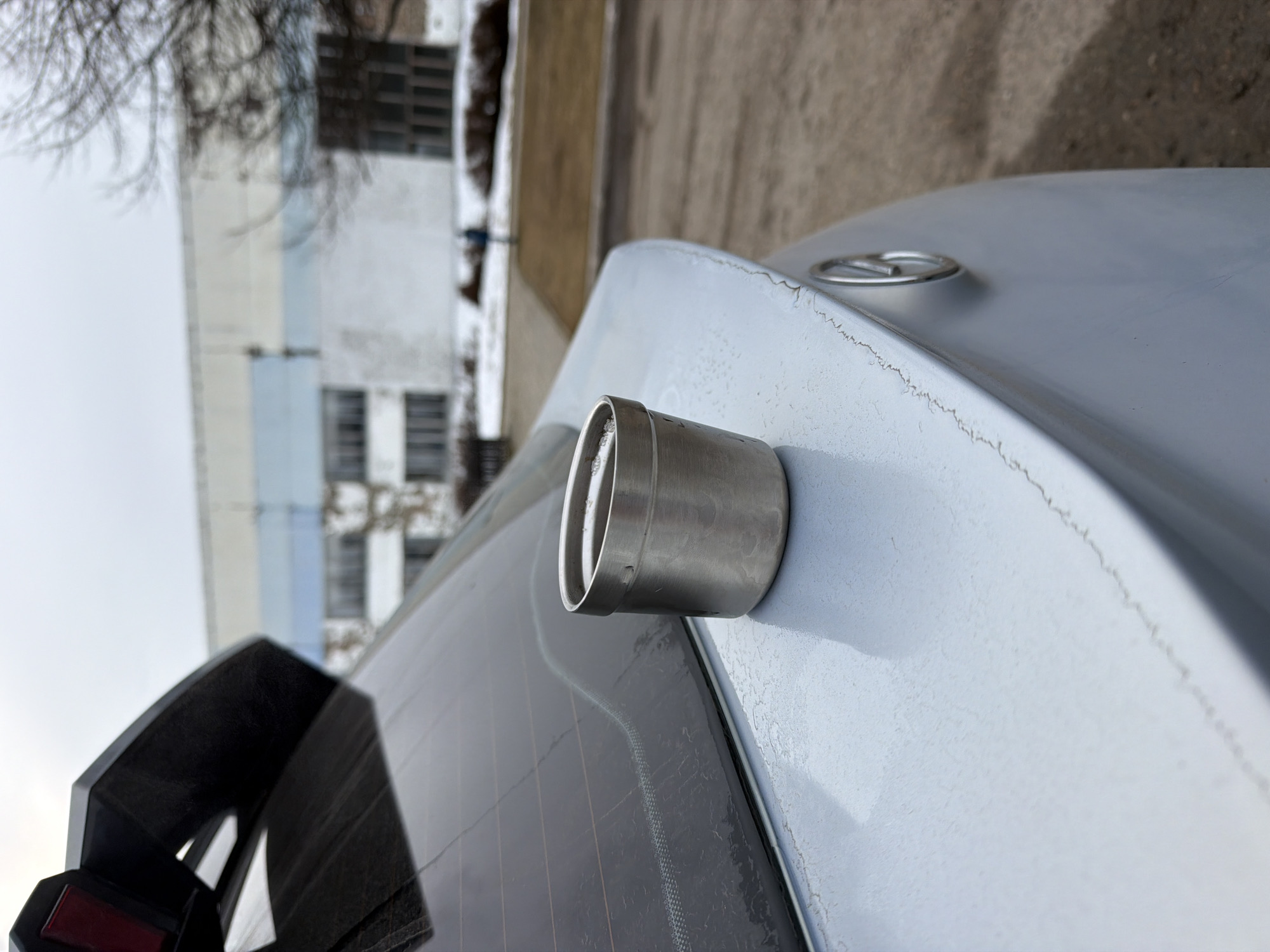 A high-angle, close-up shot focuses on the rear of a light-colored vehicle, possibly silver or grey, parked outdoors. Affixed to the trunk of the car is a cylindrical, metal object, likely an aftermarket accessory or modification. The trunk lid exhibits signs of aging, with visible cracks and imperfections in the paint. A portion of the car's rear window is visible, along with a part of a black spoiler.
In the background, slightly out of focus, are buildings. One appears to be a multi-story structure with a combination of light and dark panels, while another is a lighter-colored building with visible windows. A strip of dry, brown grass or soil is also present in the background.
Based on visual cues and general geographical knowledge, it's not possible to definitively confirm that this image was taken in Chaikovsky, Russia, based on the image alone. Additional information, such as license plates or location-specific landmarks, would be needed for confirmation.