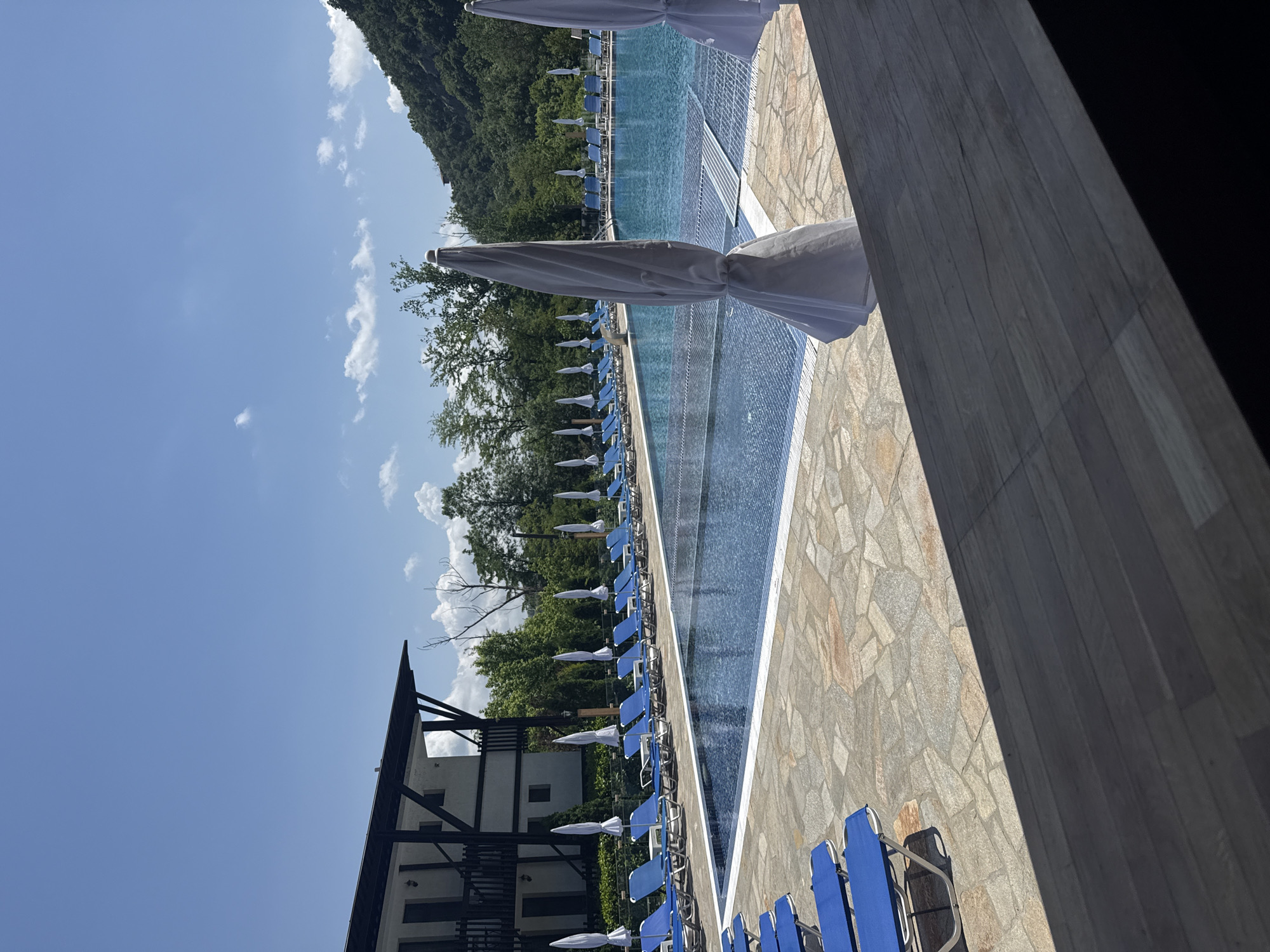The image shows a long outdoor swimming pool at a resort near Veliko Tarnovo, Bulgaria. The pool runs parallel to rows of blue lounge chairs under closed white umbrellas. Lush green trees and hills rise in the background under a blue sky with scattered clouds. A building with dark wooden trim is visible on the left side of the frame. The foreground is dominated by a textured stone patio and a dark, shadowed corner of what appears to be a building structure.