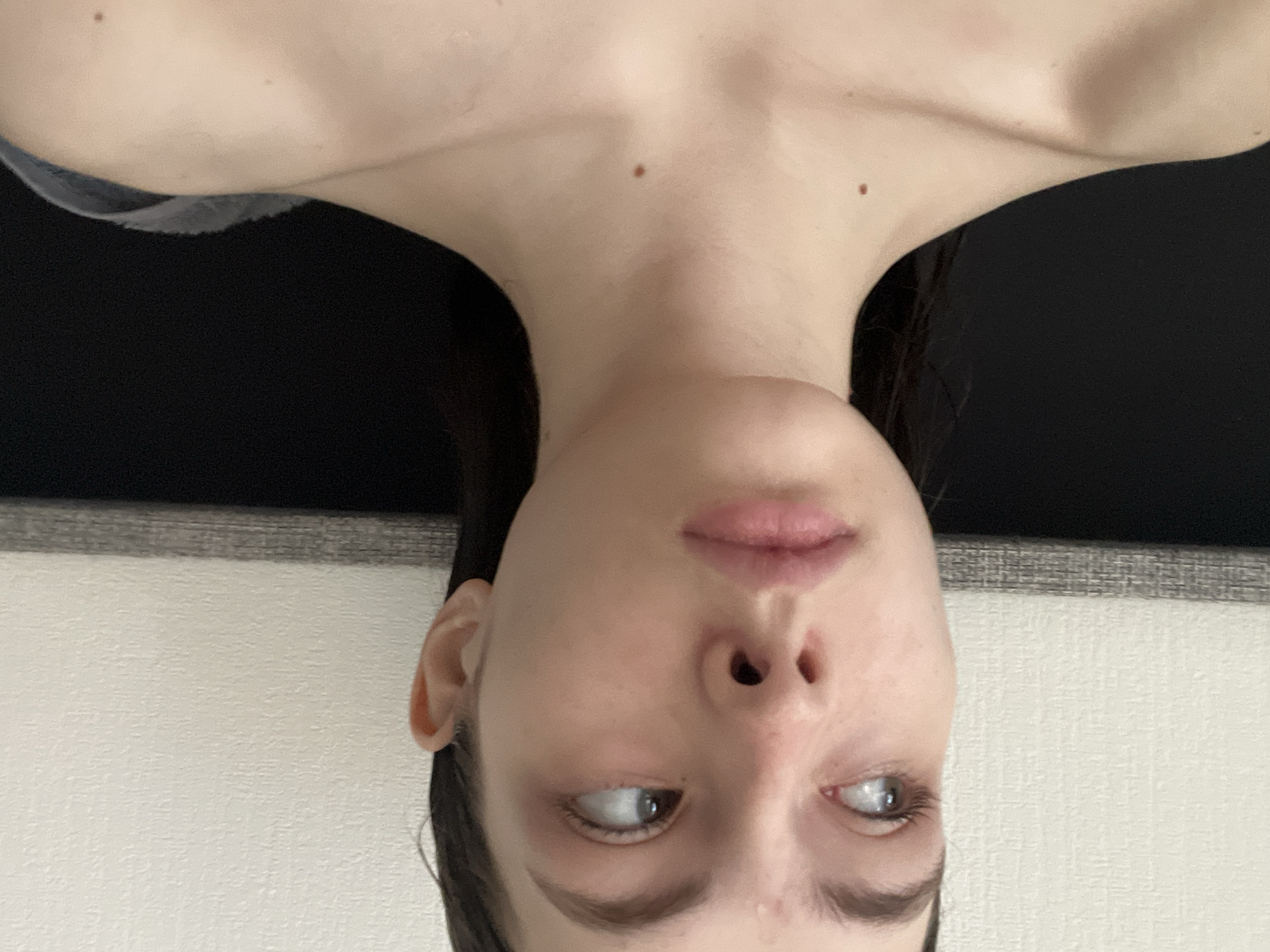 The image shows a woman's head and upper chest. She has pale skin, dark hair, and light-colored eyes. Her gaze is directed to the left. She has a small mole on her chest near her neck. She is wearing a dark-colored garment. The background consists of a light-colored wall or surface and a dark-colored horizontal area above it. The image is taken at an unusual angle, as if the camera is beneath the subject. There is no evidence to suggest it was taken in Saint Petersburg, Russia.