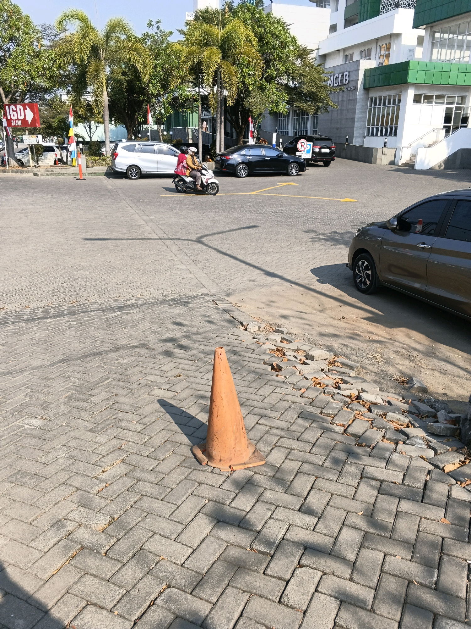 The image depicts an outdoor scene in Semarang, Indonesia, likely in front of a building identified by the signage "MCE B". The foreground is dominated by a gray, herringbone-patterned paver surface with a visible orange traffic cone. To the right, the rear section of a dark grey car is parked. In the mid-ground, a person on a scooter, wearing a helmet and a light-colored jacket over a darker top, is seen driving towards the left. Behind the scooter, a silver car is parked. Further back, a black SUV is visible, with a circular "no parking" sign attached to its rear. The background features palm trees and a modern building with green and white accents. A red and white flag is also present. The overall environment appears to be a paved area, possibly a parking lot or a street, adjacent to a building. The sky is clear and blue, suggesting daytime.